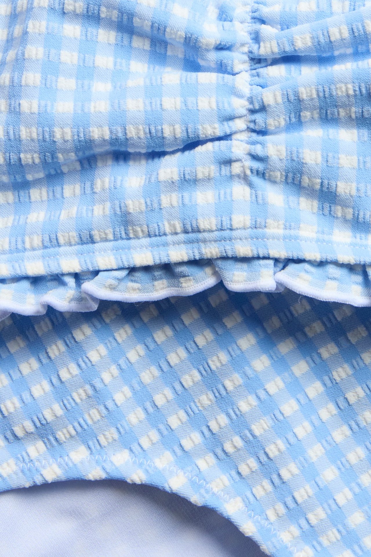 Zbliżenie na niebiesko-białe, kraciaste bikini dziecięce z seersuckeru, z marszczeniem i falbanką.