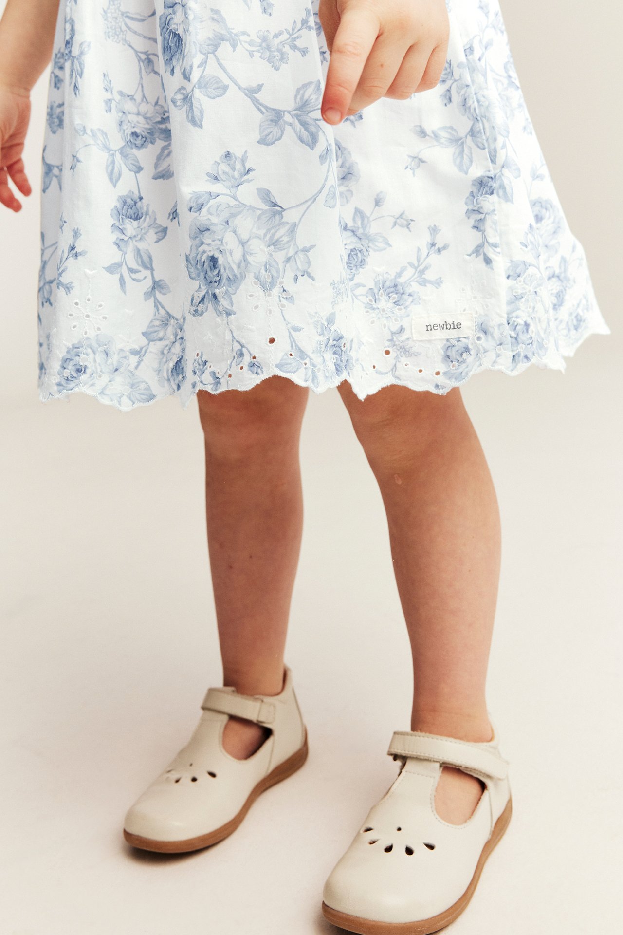 Barn i off-white blomstret vevd kjole med blå blomster, brodert kant og beige Mary Jane sko, forfra.