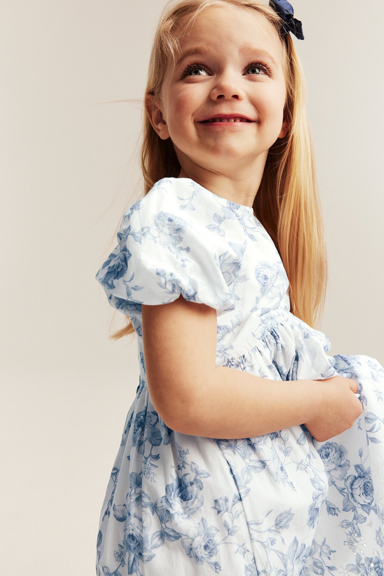 Nærbilde av en jente i en offwhite blomstret kjole med puffermer og blå h
R
Kid-jente i offwhite blomstret kjole med puffermer og blå h
R
Kid-jente i offwhite blomstret kjole med puffermer og blå hårsløyfe.