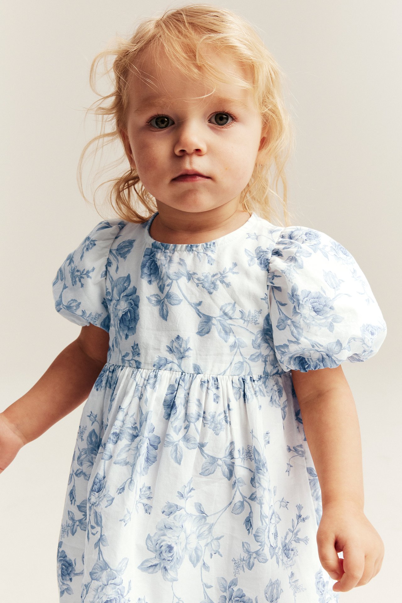 Frontvy av ett barn i offwhite klänning med blått blommönster och puffärmar.