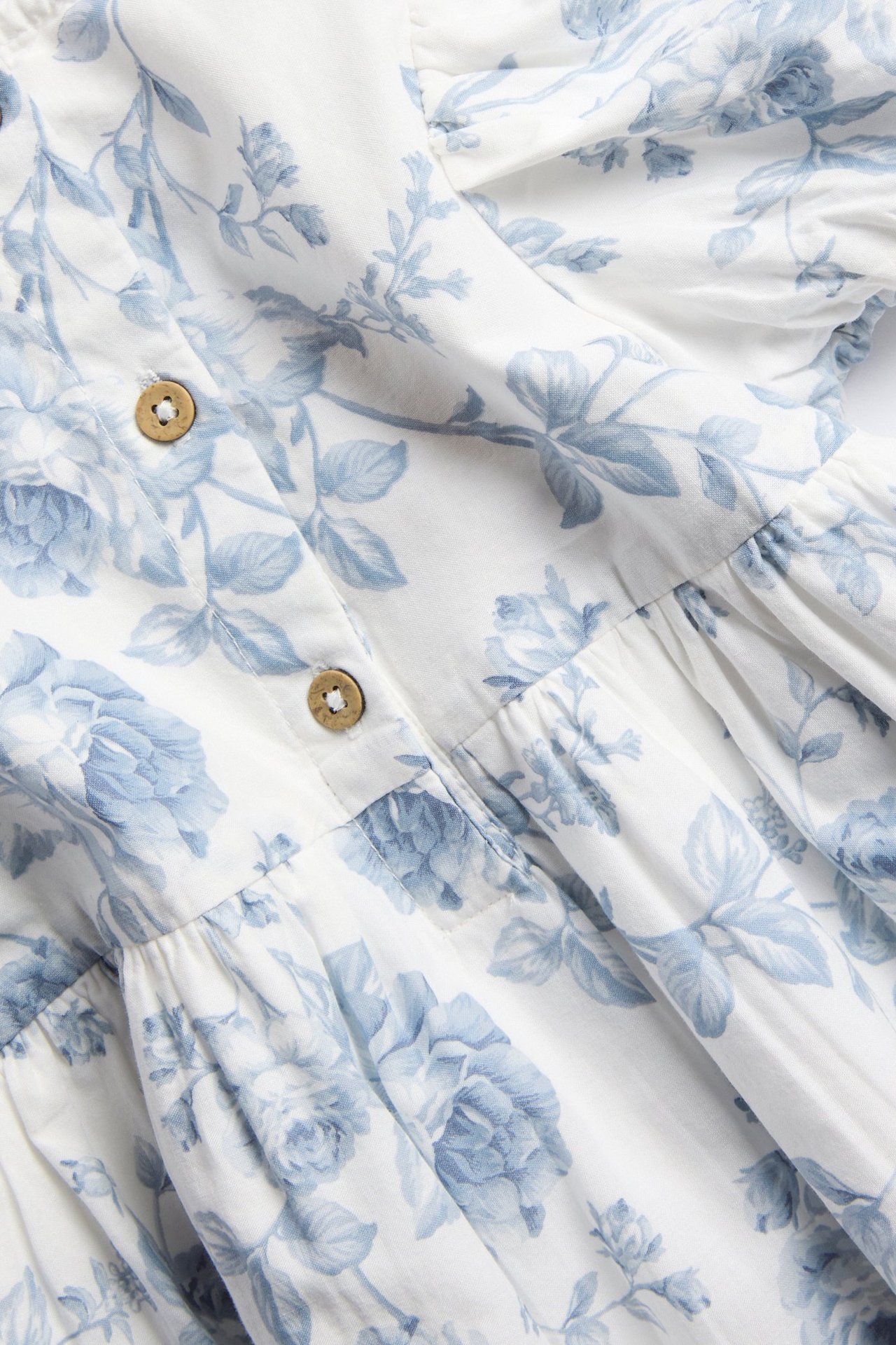 Nærbilde av en offwhite damekjole med lysblått blomstermønster, som viser to treknapper og rynket stoff.