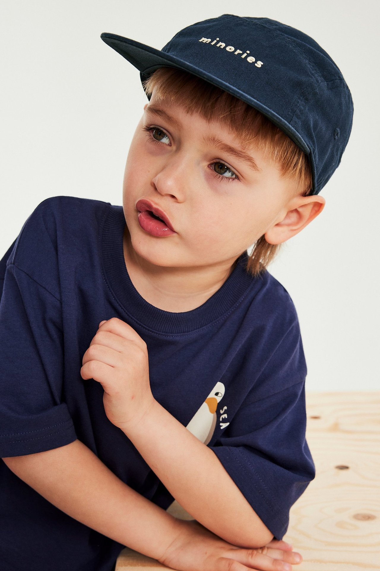 Edestäpäin: lapsi tummansinisessä puuvillalippalakissa valkoisella "minorities"-kirjailulla ja tummansinisessä t-paidassa.