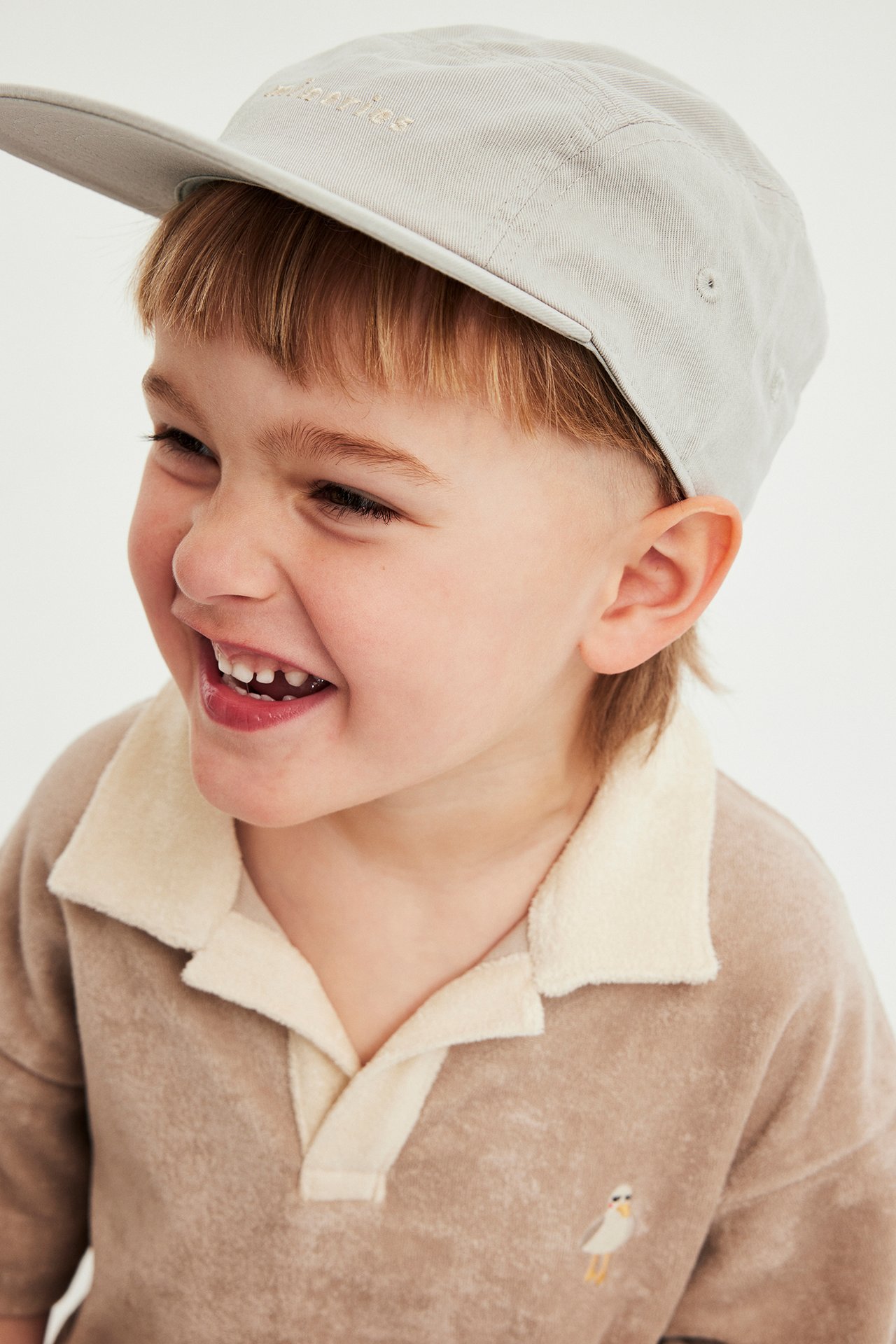 Sivukuva, hymyilevä lapsi yllään beige lippalakki brodeerauksella ja säädettävällä takaosalla, sekä ruskea froteepaita.