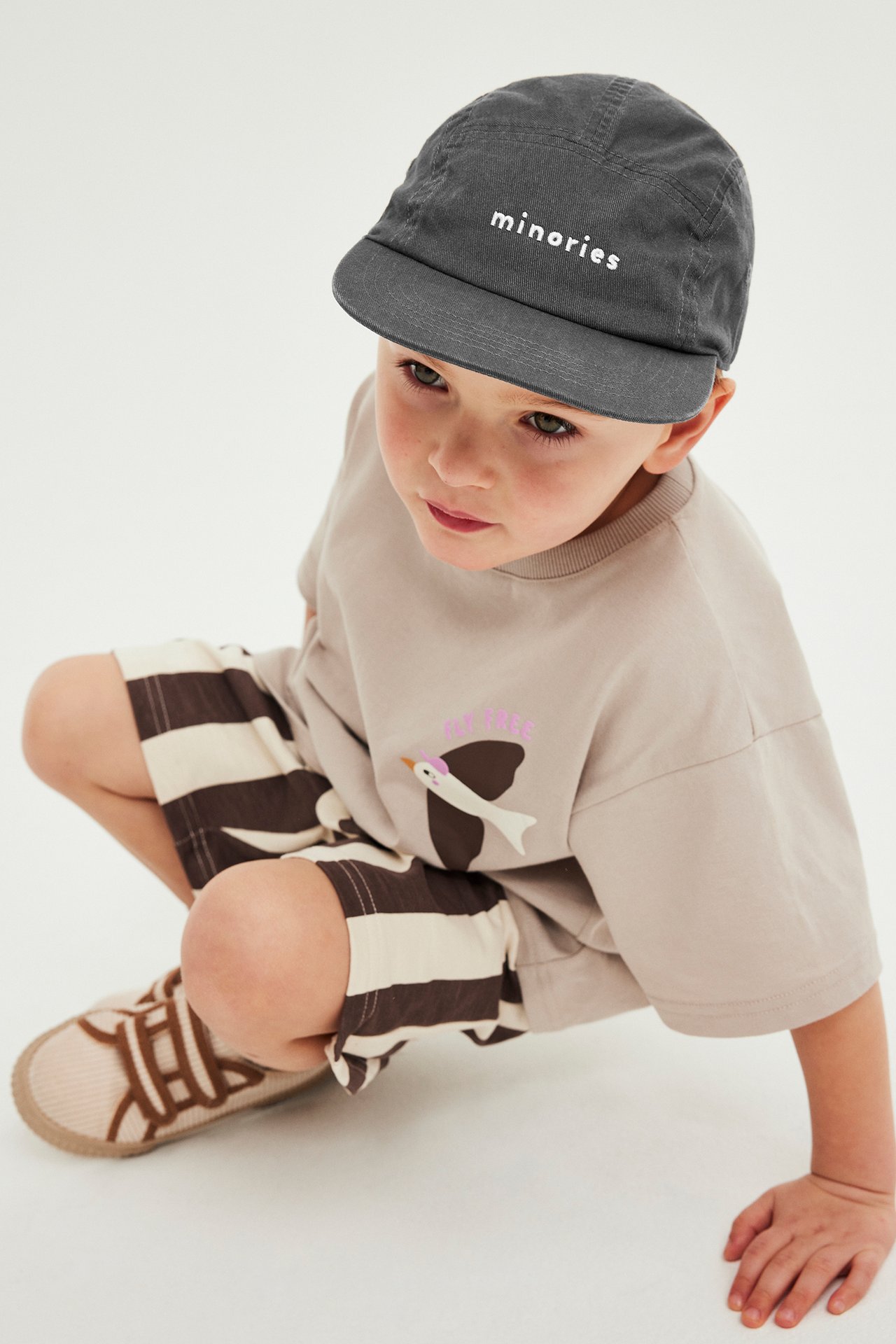 Kohotettu etunäkymä: Lapsi mustassa brodeeratussa puuvillakepsissä, beigessä t-paidassa, ruskeajuovaisissa shortseissa ja beigissä tennareissa.