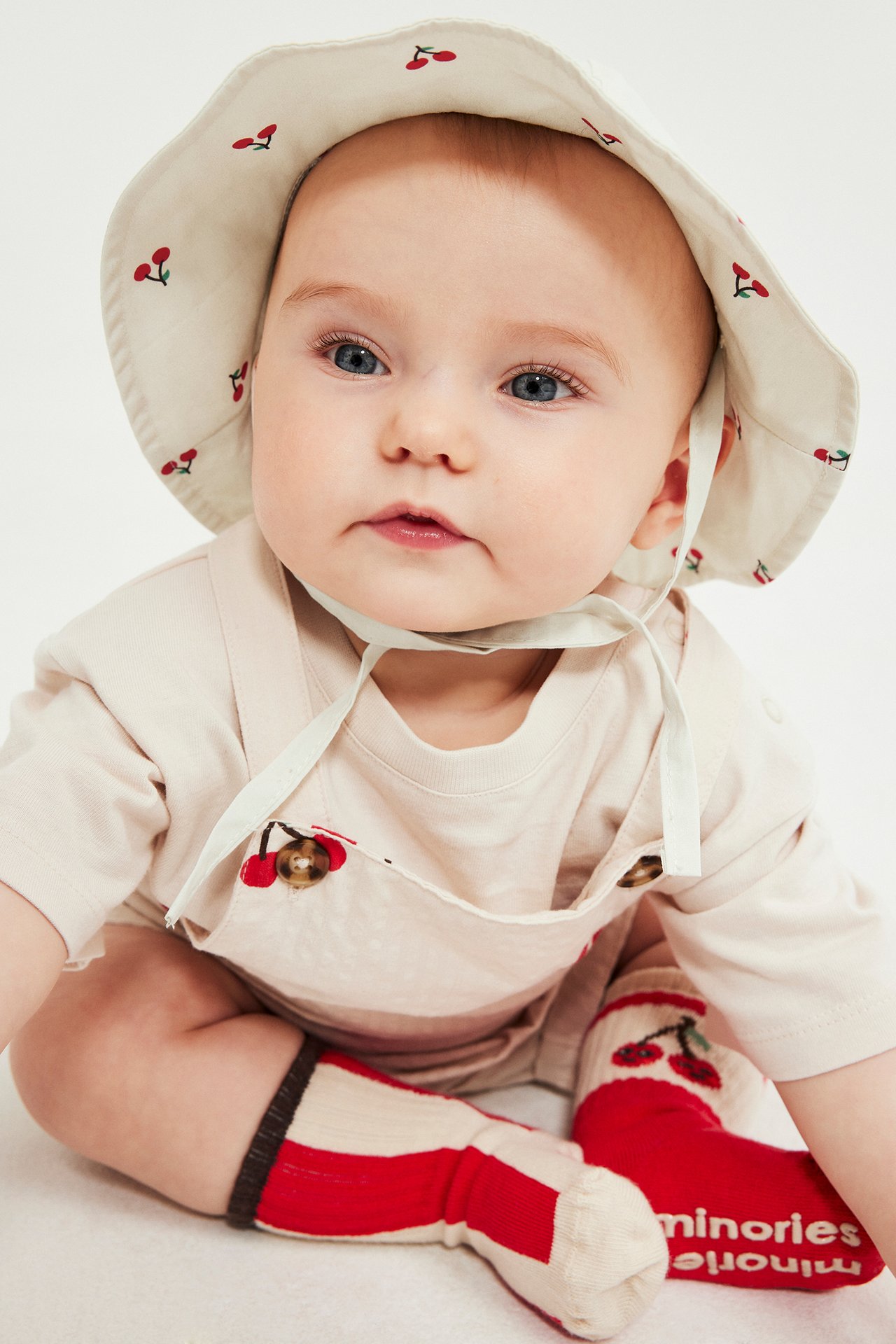 Frontvy av baby i beige bomulls solhatt med kirsebærmønster, beige snekkerbukse og rødstripete sokker.