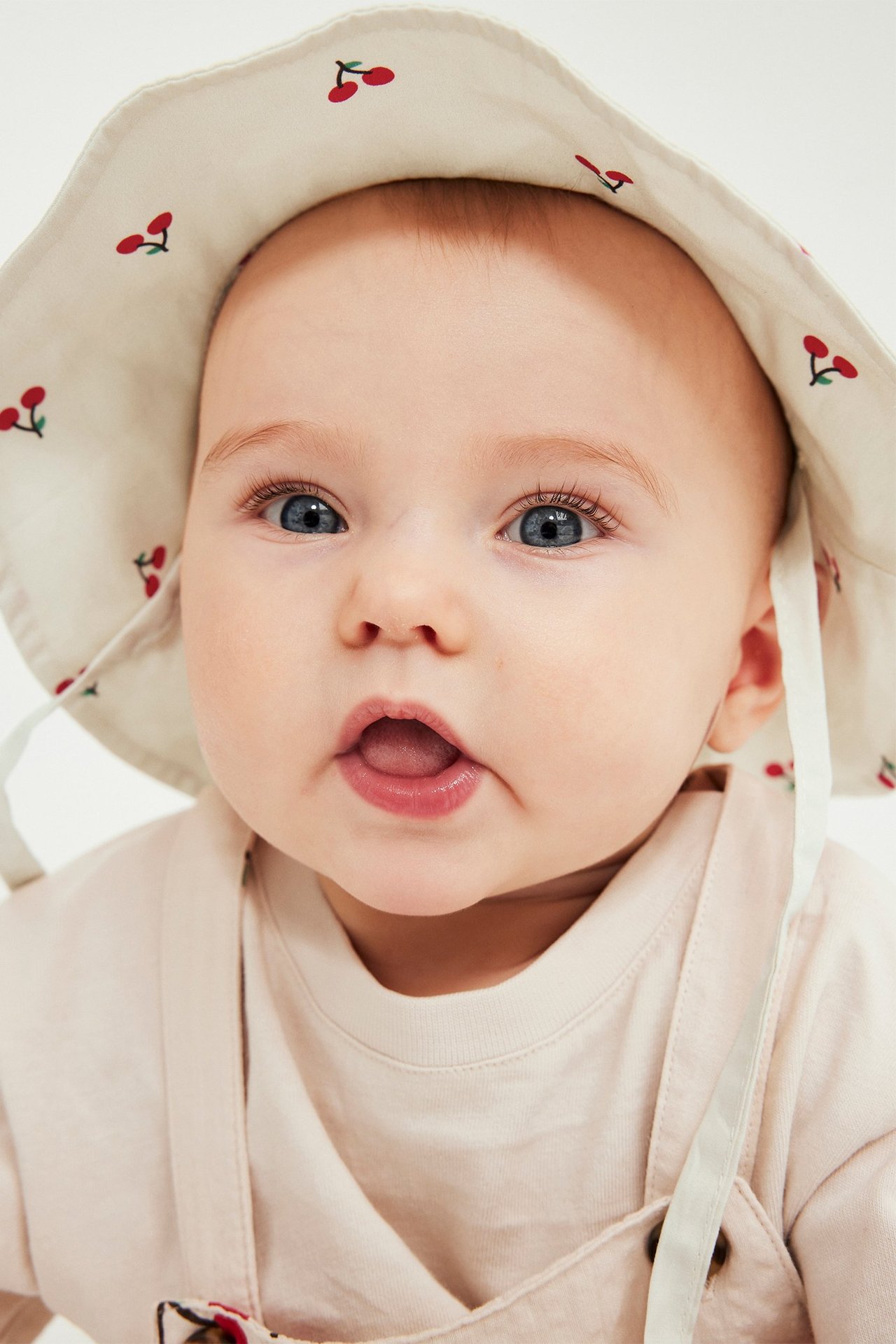 Zbliżenie przodem: niemowlę nosi beżowy bawełniany kapelusz przeciwsłoneczny w wiśnie z troczkami, na jasnym topie.