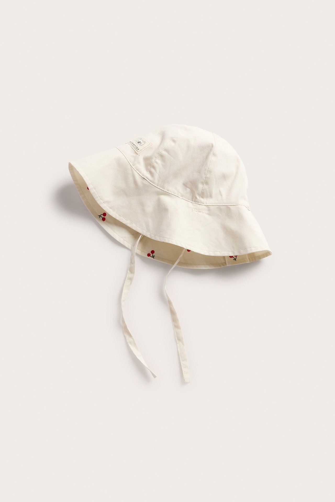 Hög vinkelvy av en beige solhatt i bomull för baby, med knytband och körsbärsmönstrat foder.
