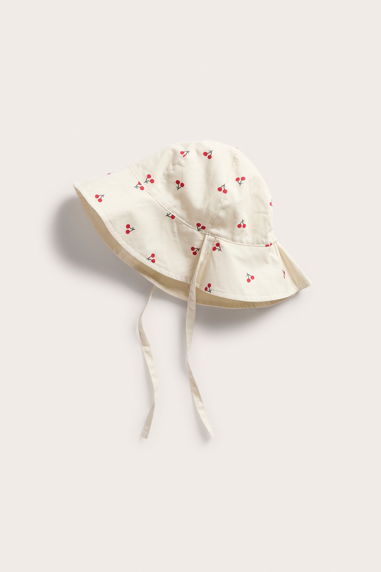 Beige solhatt i bomull för baby med rött körsbärsmönster och knytband, sett framifrån.
