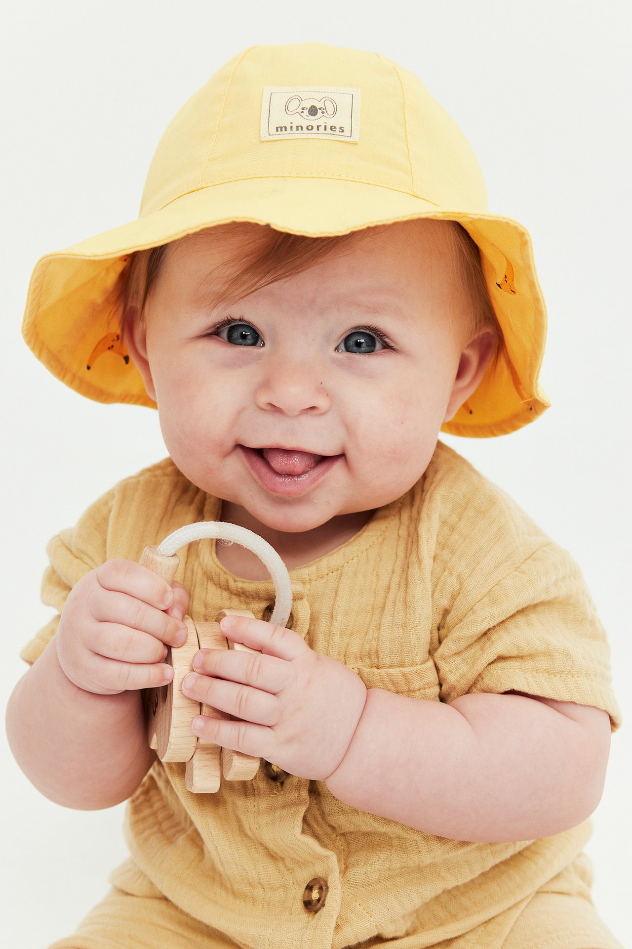 Edestä: Baby keltaisessa käännettävässä aurinkohatussa banaaniprintillä, keltaisessa haalarissa, pitää puulelua.