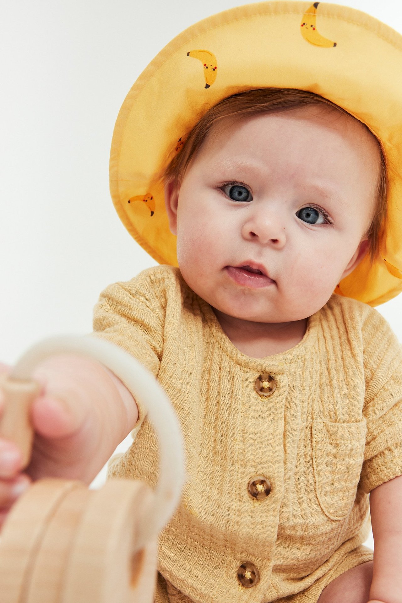 Lähikuva vauvasta keltaisessa banaanikuvioisessa aurinkohatussa ja keltaisessa puuvillabodyssa, pitäen puista lelua.