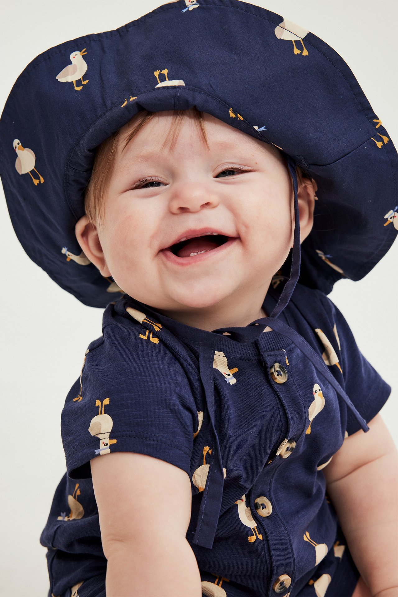 Närbild framifrån på en leende baby i mörkblå solhatt med ank-mönster och matchande body.