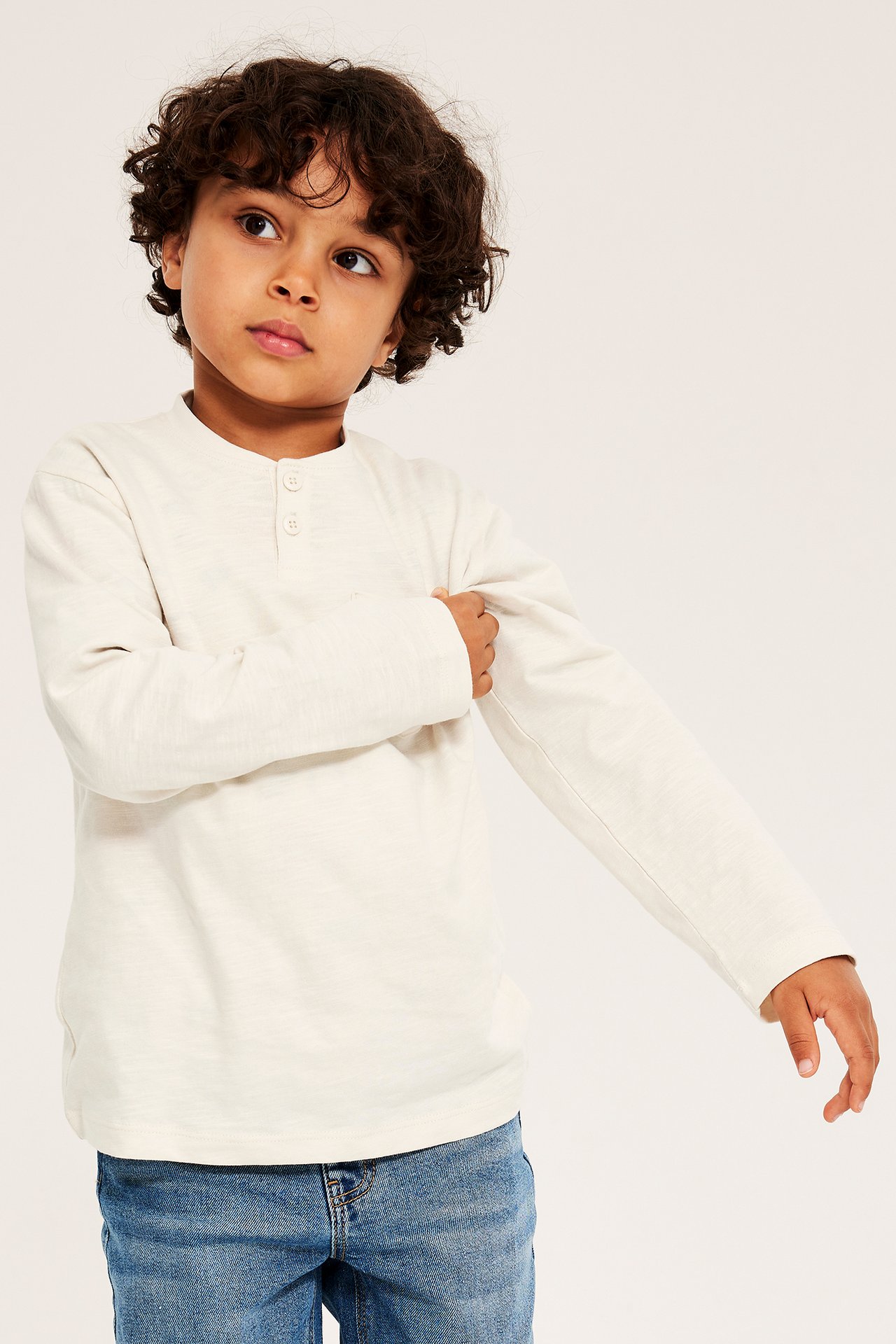 Framifrån, ett barn i ljusbeige bomulls-farfarströja med knappar, bröstficka och blå jeans.