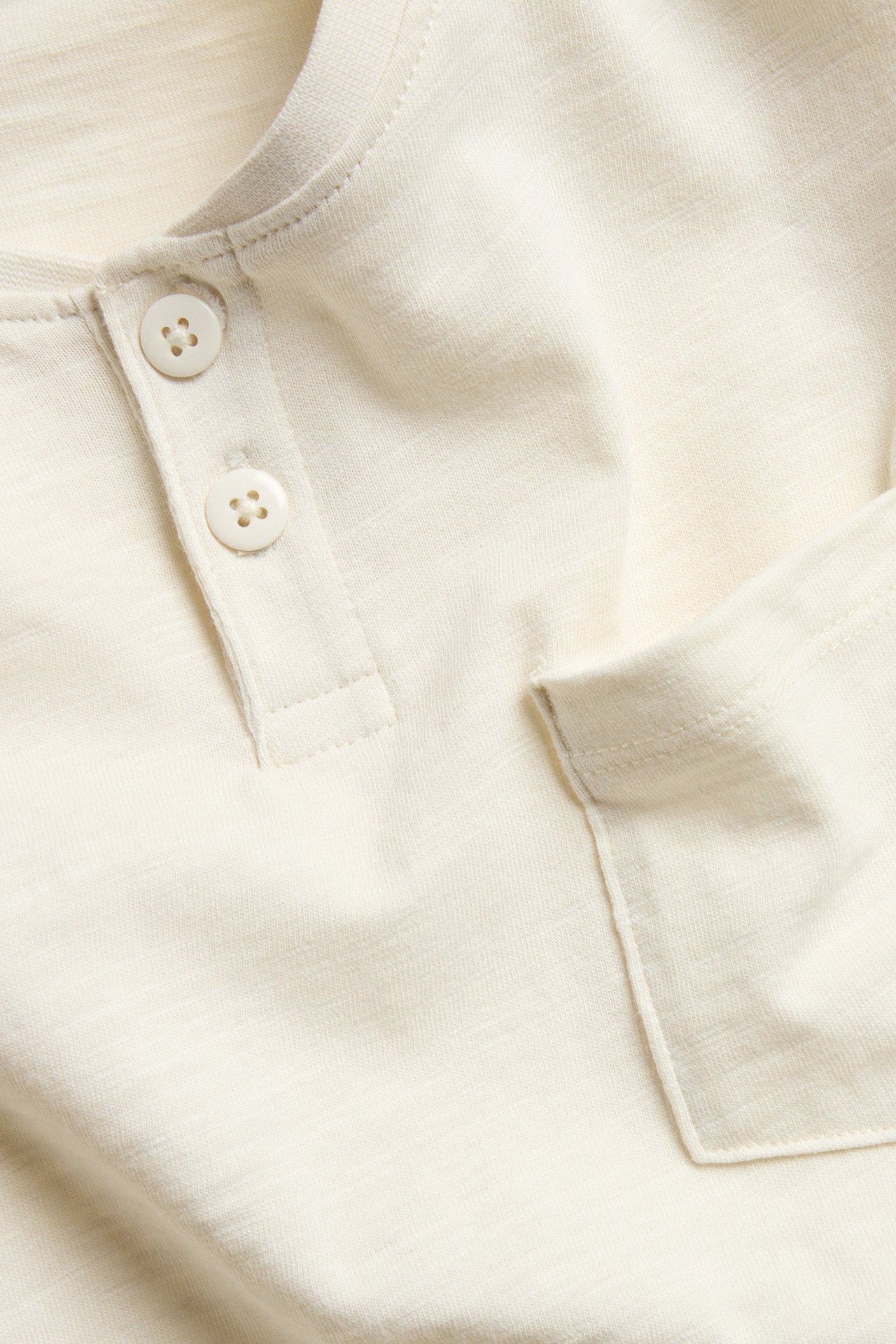 Zbliżenie na jasnobeżową koszulkę Henley z bawełny organicznej ze splotem slub, z dwoma guzikami i kieszenią.
