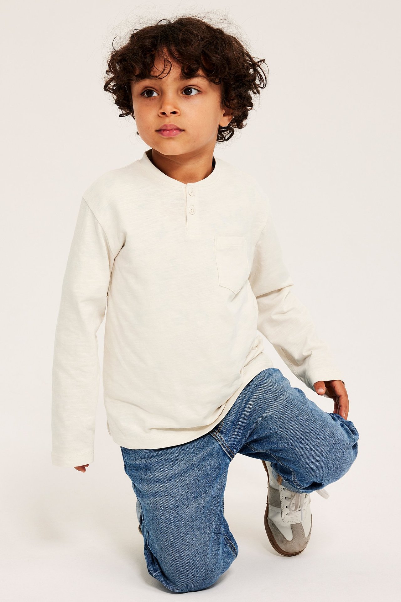 Frontvy av ett barn i en ljusbeige bomullsfarfarströja med knappar, blå jeans och sneakers.