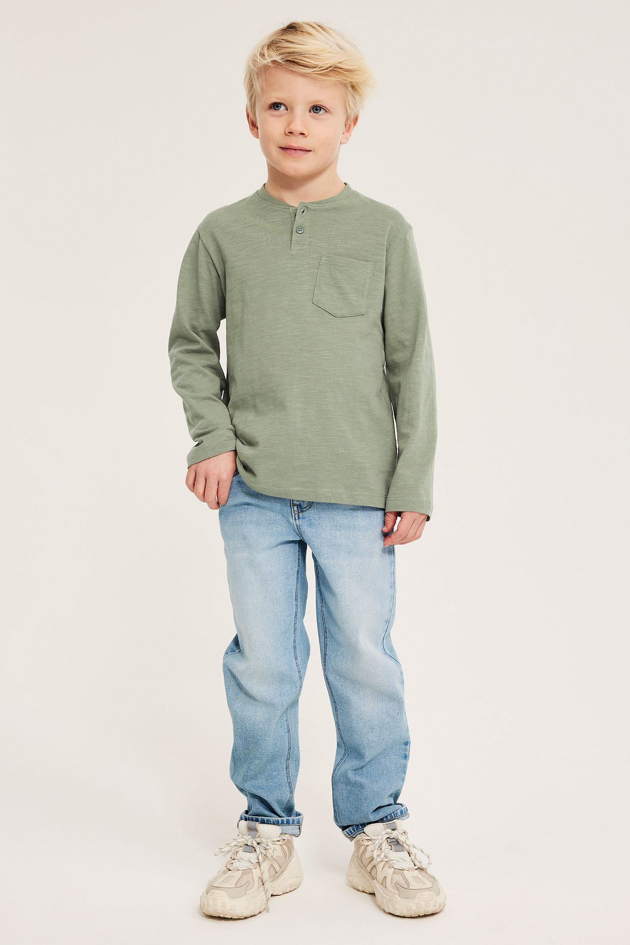 Widok z przodu: dziecko w jasnozielonej bawełnianej koszulce Henley z guzikami, jasnoniebieskich jeansach i beżowych trampkach.
