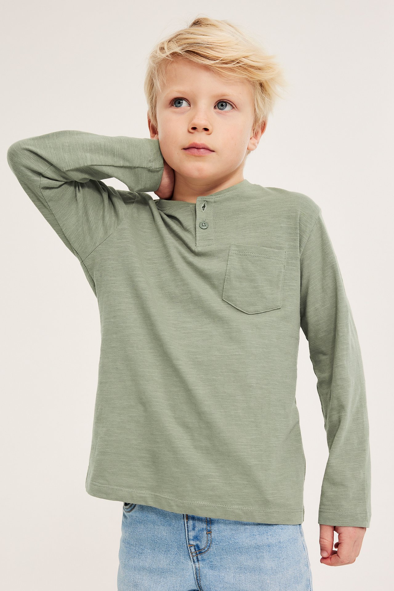 Widok z przodu: Dziecko w zielonej koszulce Henley z guzikami i jasnoniebieskich dżinsach.