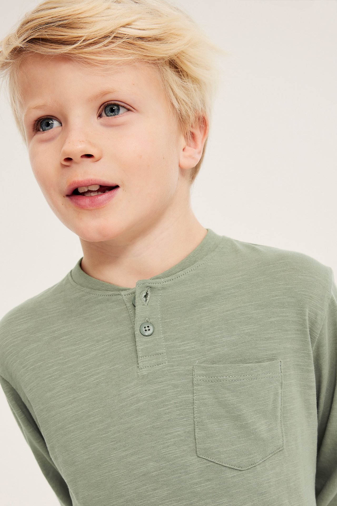 Widok z przodu: Dziecko w zakurzonej zielonej koszulce Henley z długim rękawem, guzikami i kieszonką.