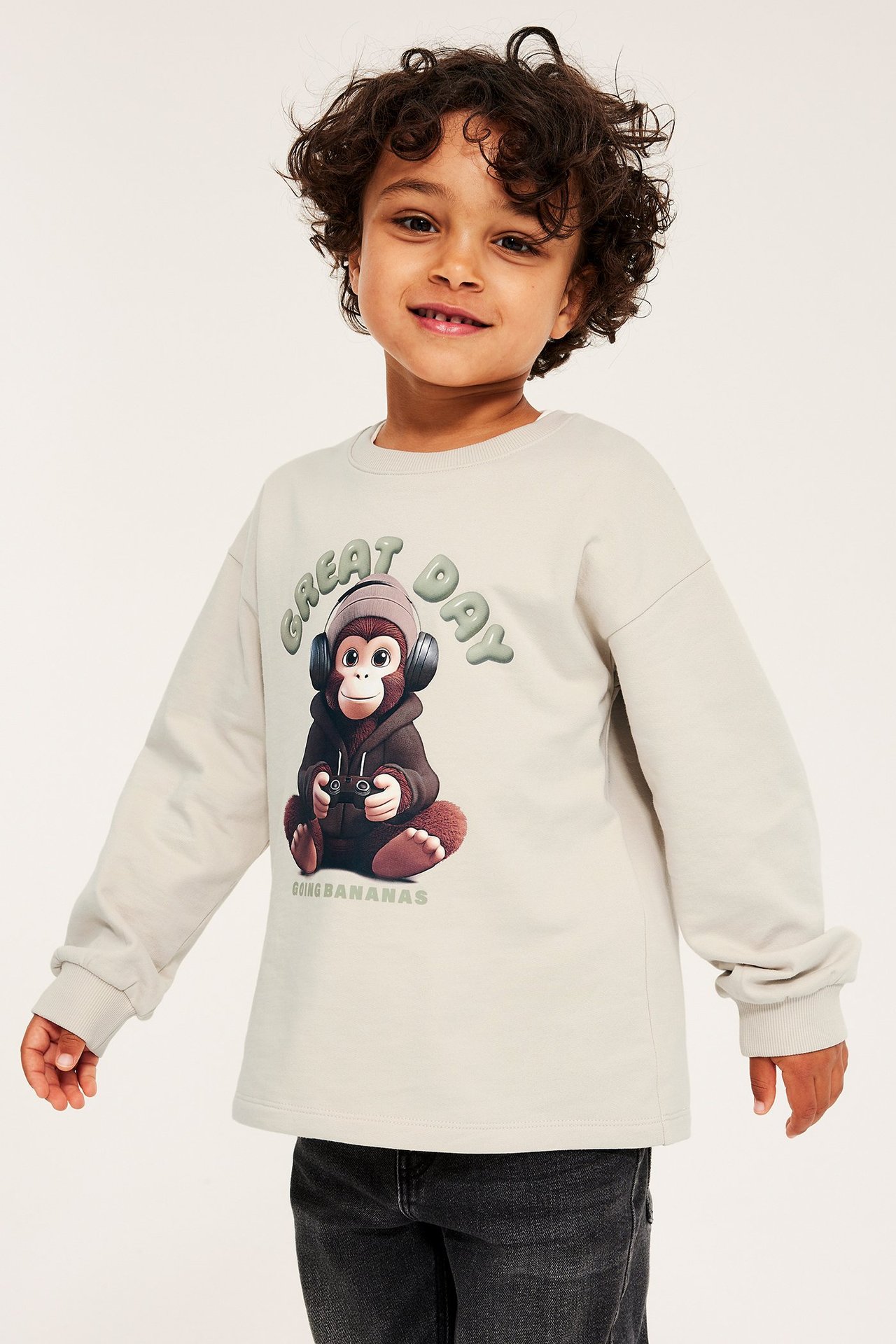 Widok z przodu dziecka w beżowej bawełnianej bluzie z nadrukiem małpy i ciemnoszarych jeansach.