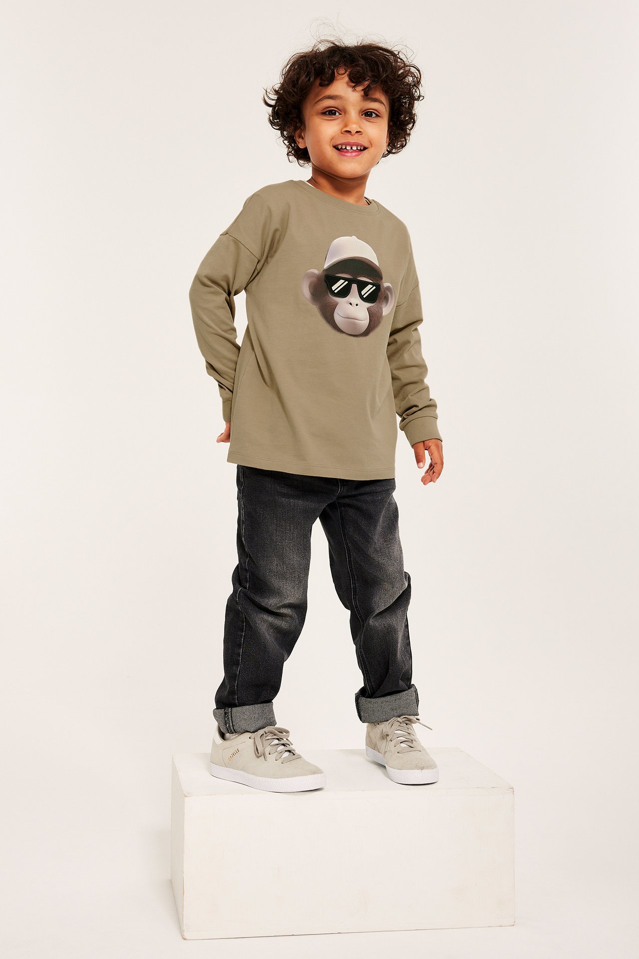 Framsida: leende barn i en beige långärmad t-shirt med ap-tryck, mörka jeans och ljusa sneakers.