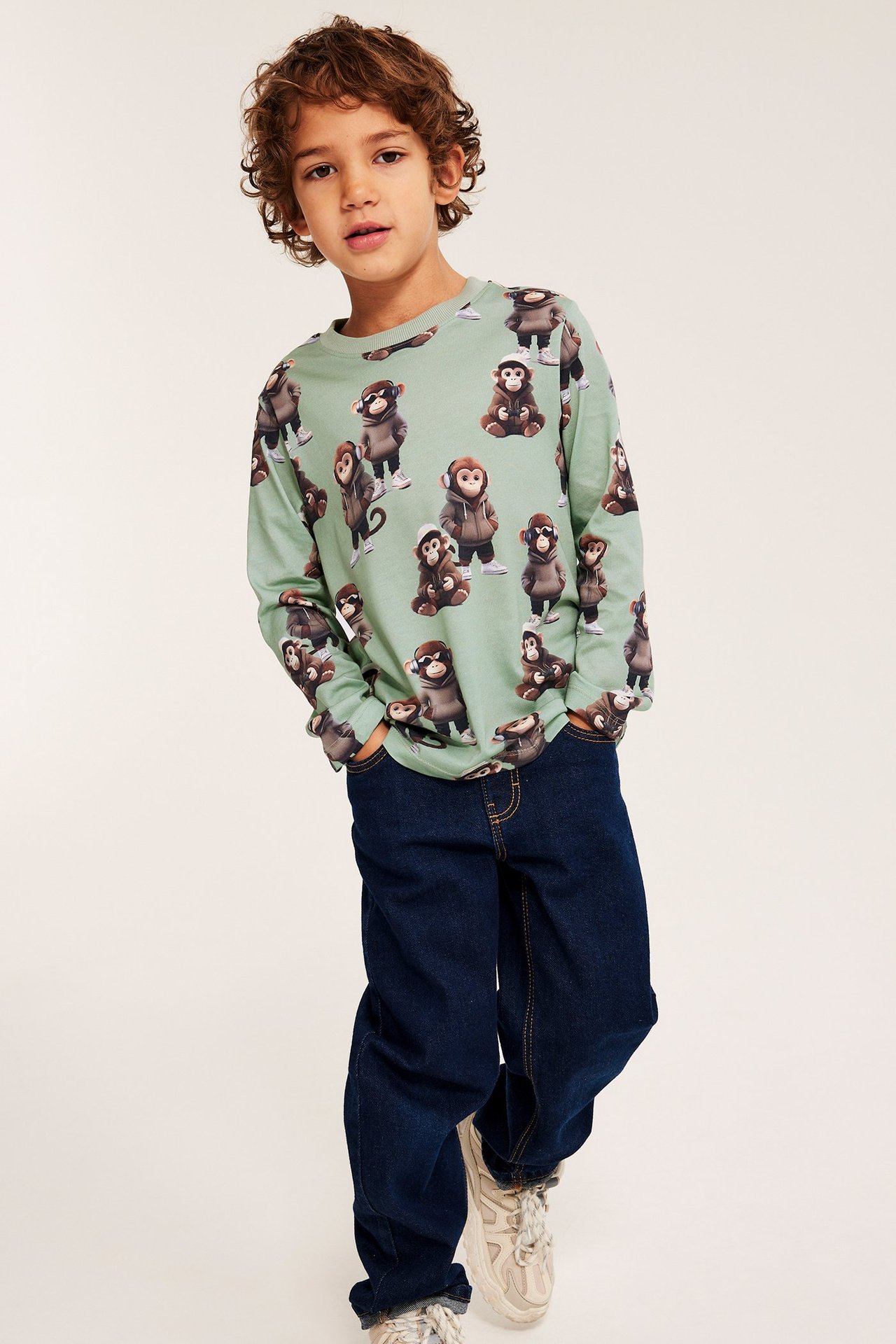 Edestä: lapsi vaaleanvihreässä apinakuvioisessa pitkähihaisessa t-paidassa, tummansinisissä farkuissa ja beigessä tennareissa.
