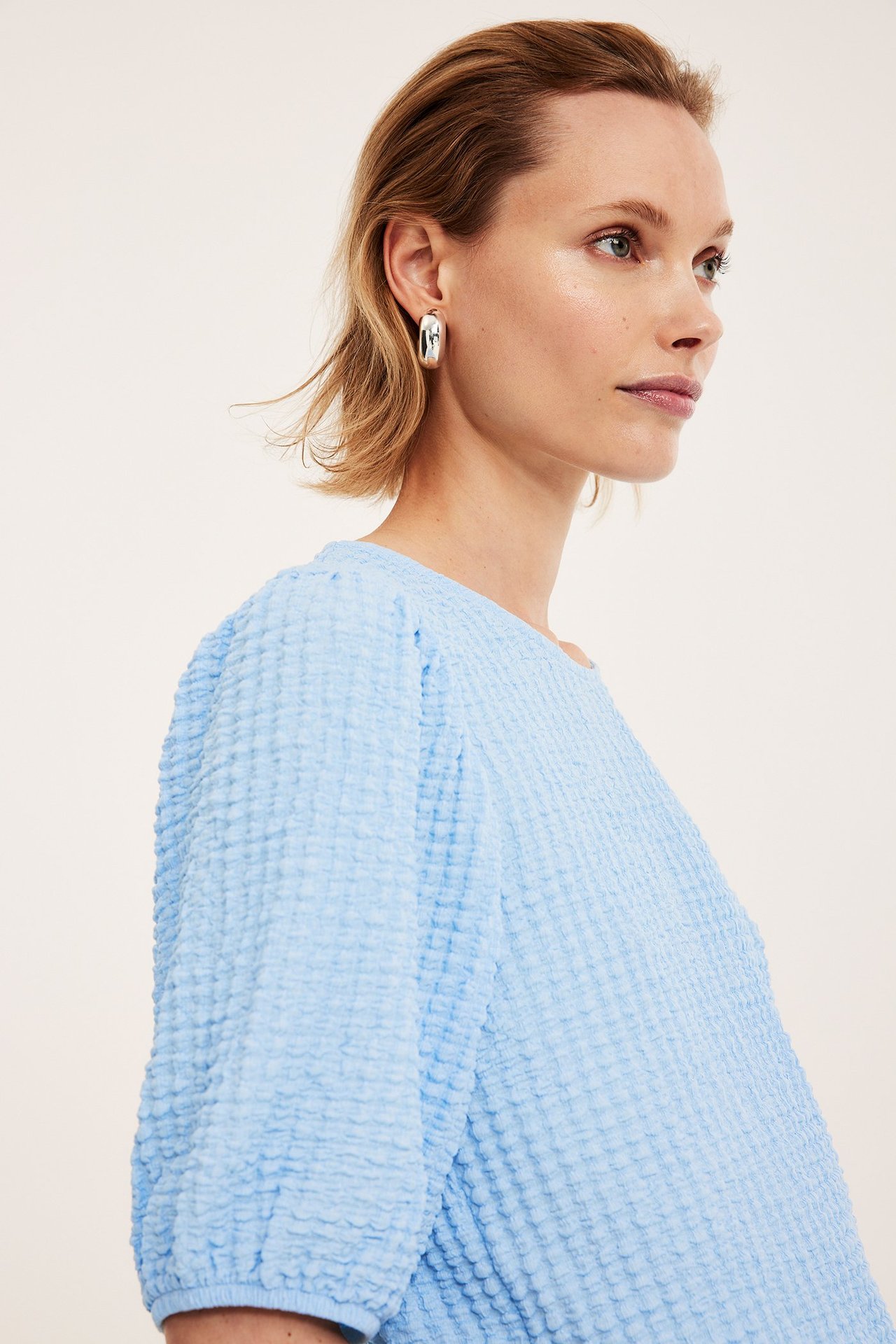 Zbliżenie, widok z boku kobiety w jasnoniebieskim topie z bufiastymi rękawami i srebrnych, grubych kolczykach kołach.