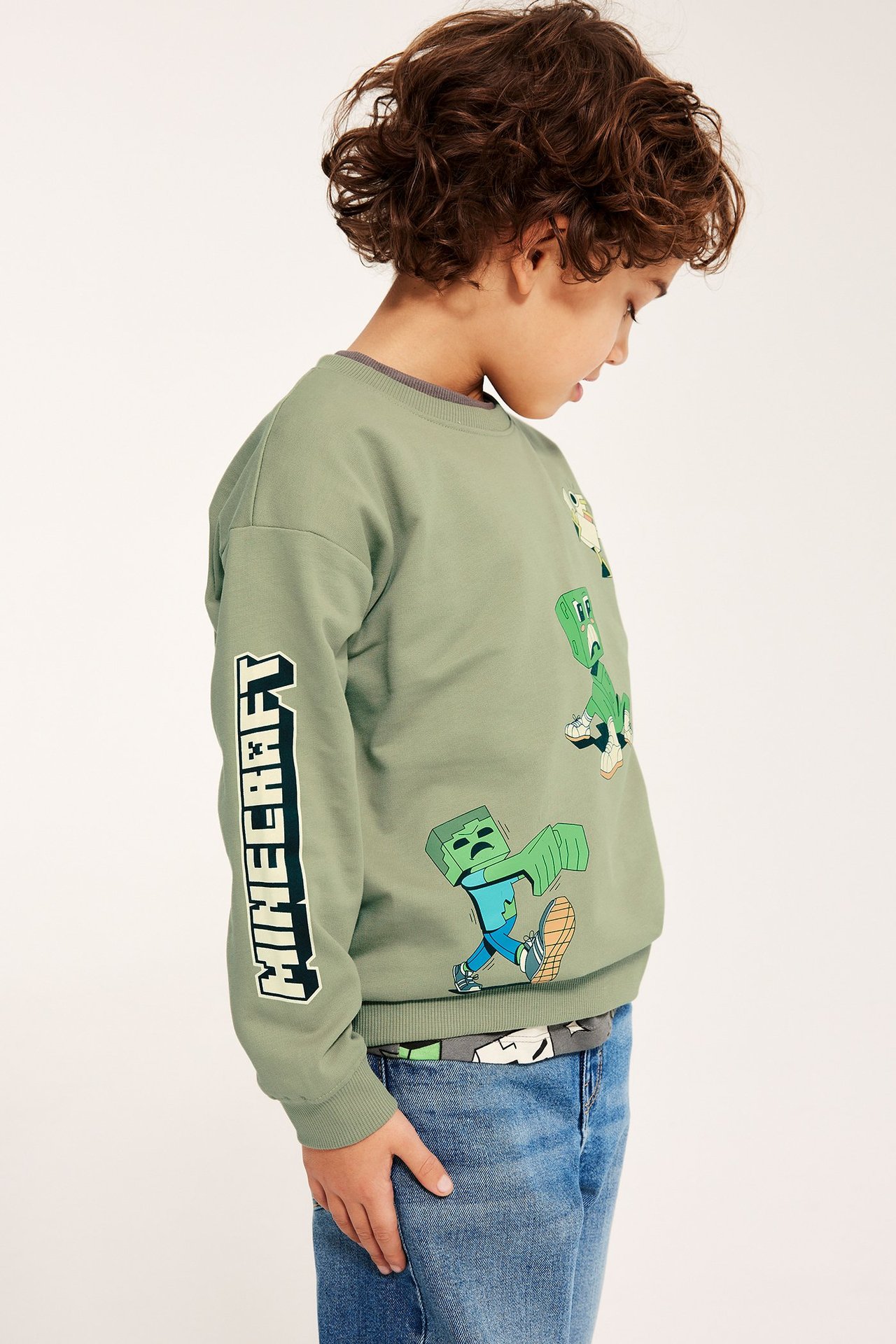 Sivukuva: Lapsi tummanvihreä Minecraft-collegepaita painatuksella, harmaan paidan ja sinisten farkkujen päällä.