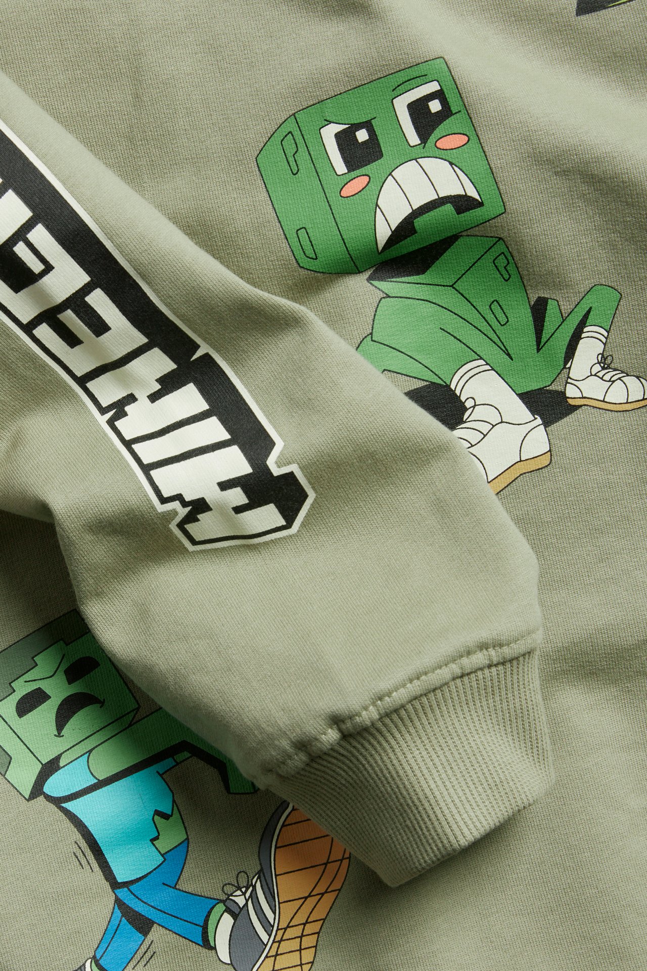 Lähikuva tummanvihreästä lasten puuvillapaidasta, jossa on Minecraft-hahmo- ja logopainatus hihassa.