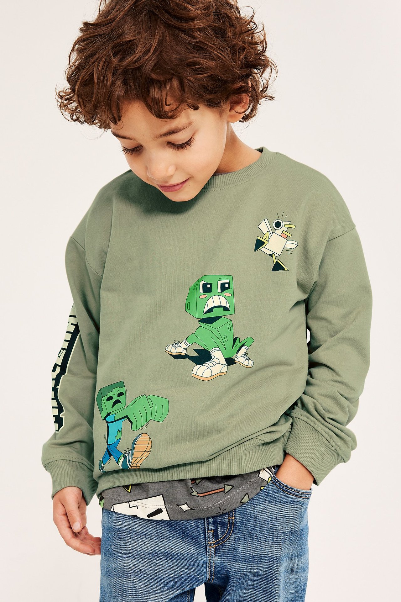 Etukuva lapsesta yllään tummanvihreä puuvillainen Minecraft-collegepaita kuvaprinteillä, sinisten farkkujen kanssa.