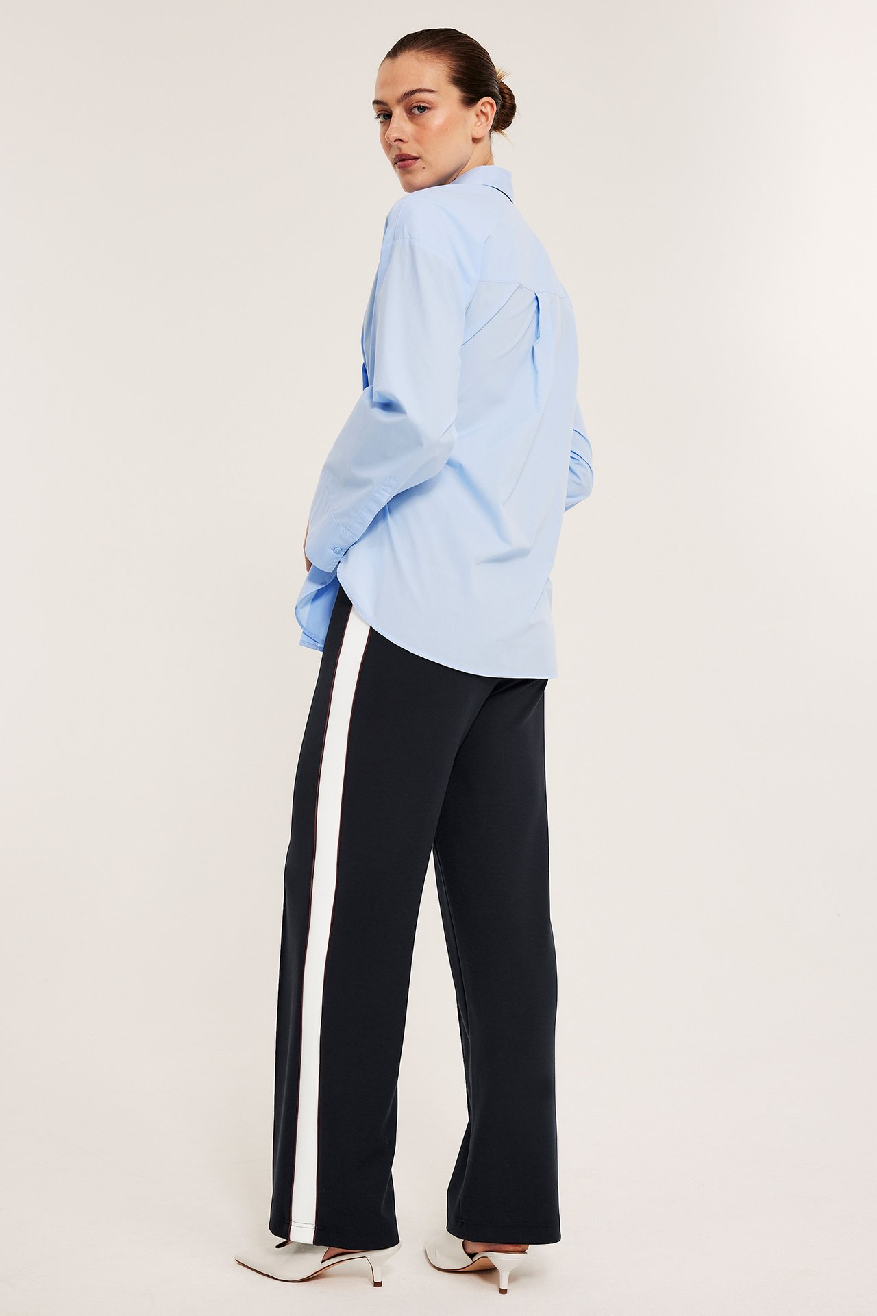 Kvinne i mørkeblå vide jerseybukser med hvite sidestriper, lyseblå skjorte og hvite hæler.