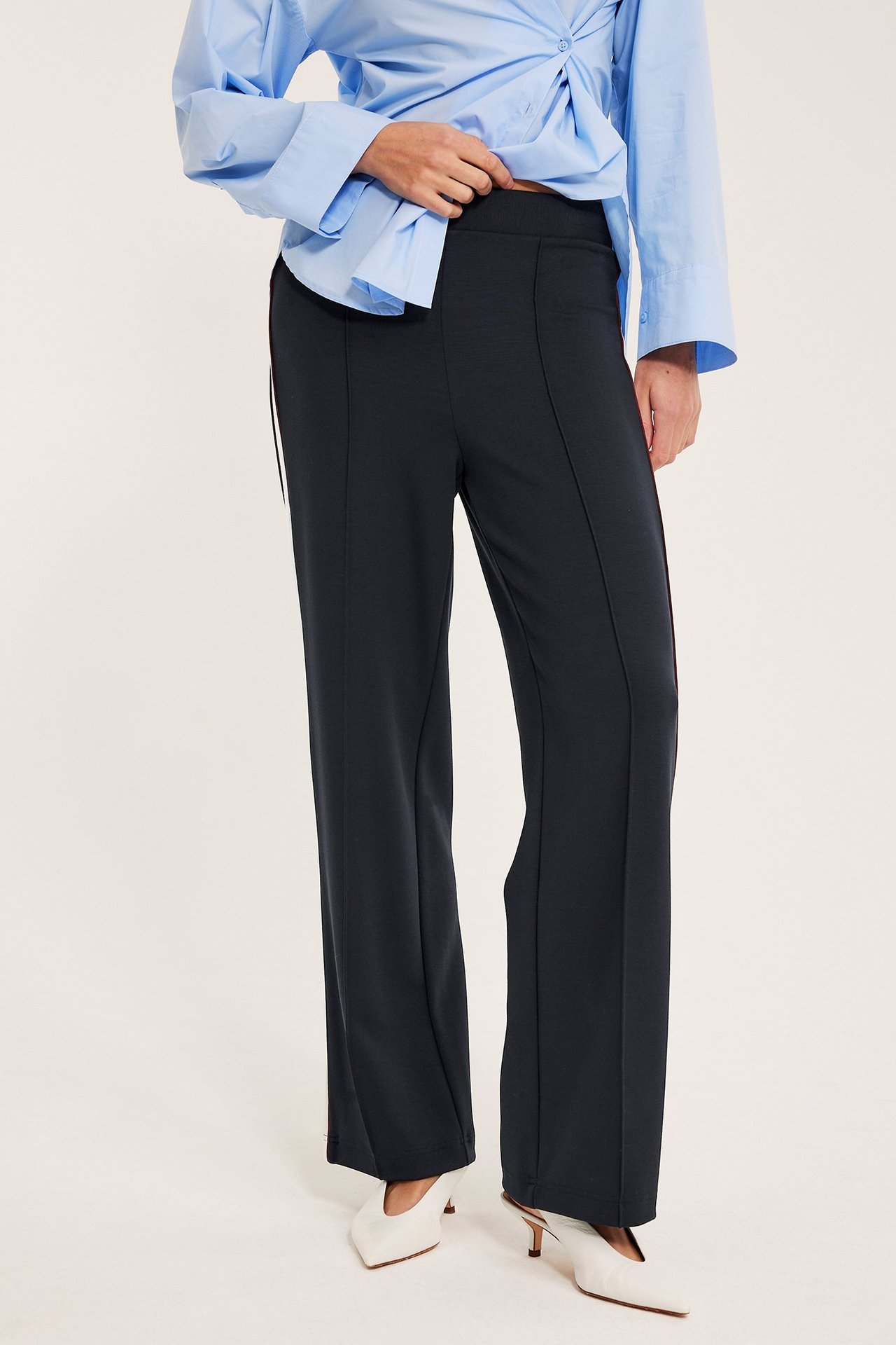 Nainen pukeutuneena tummansinisiin leveisiin jerseyhousuihin, joissa on prässit ja raita, siniseen paitaan ja valkoisiin korkokenkiin.