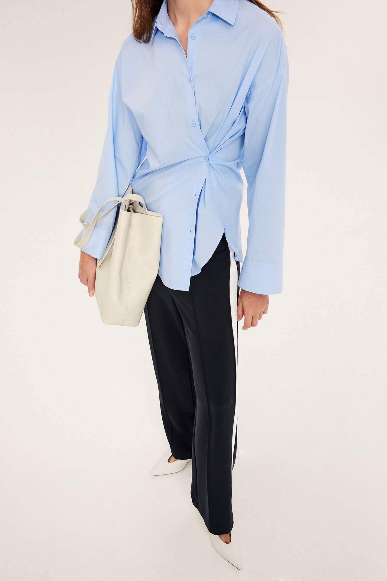 Kvinna i mörkblå vida jerseymjukisbyxor med vit sidorand, ljusblå tvinnad skjorta, vita skor, håller beige tygväska.