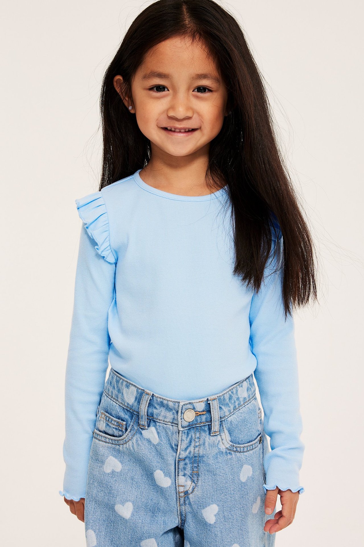 Framsida: Ett barn i ljusblå ribbad långärmad topp med volanger, matchad med hjärtmönstrade jeans.