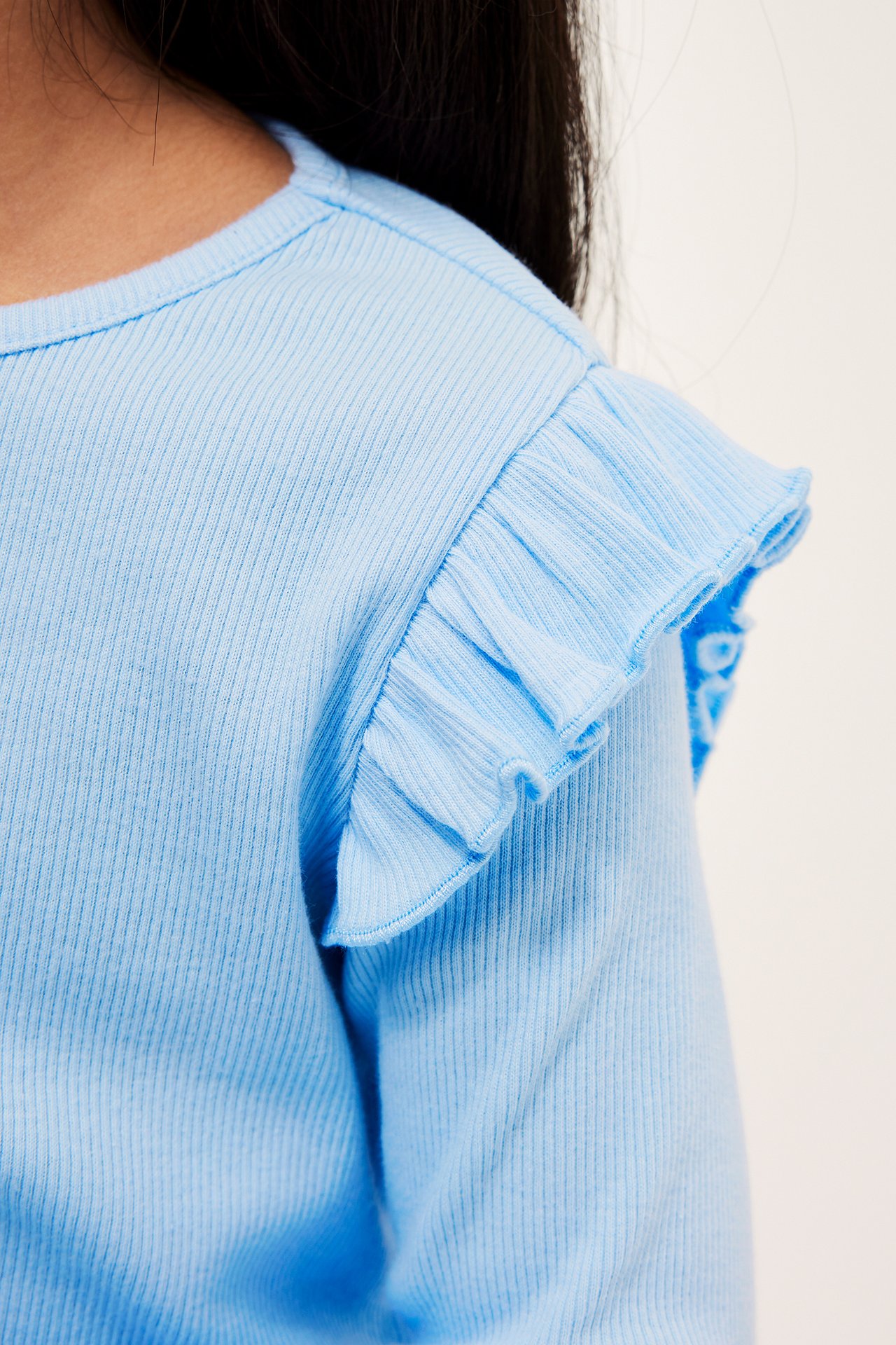 Nærbilde av en lyseblå ribbestrikket langermet topp for barn med rysjer på skuldrene.