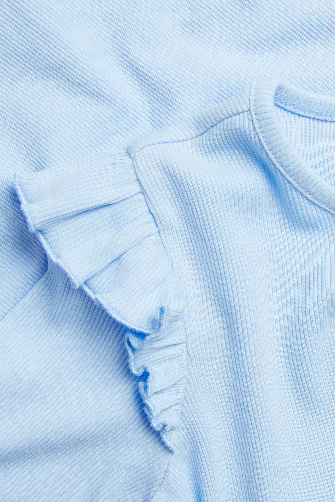 Nærbilde av en lyseblå ribbestrikket langermet topp med volangdetalj på skulderen.