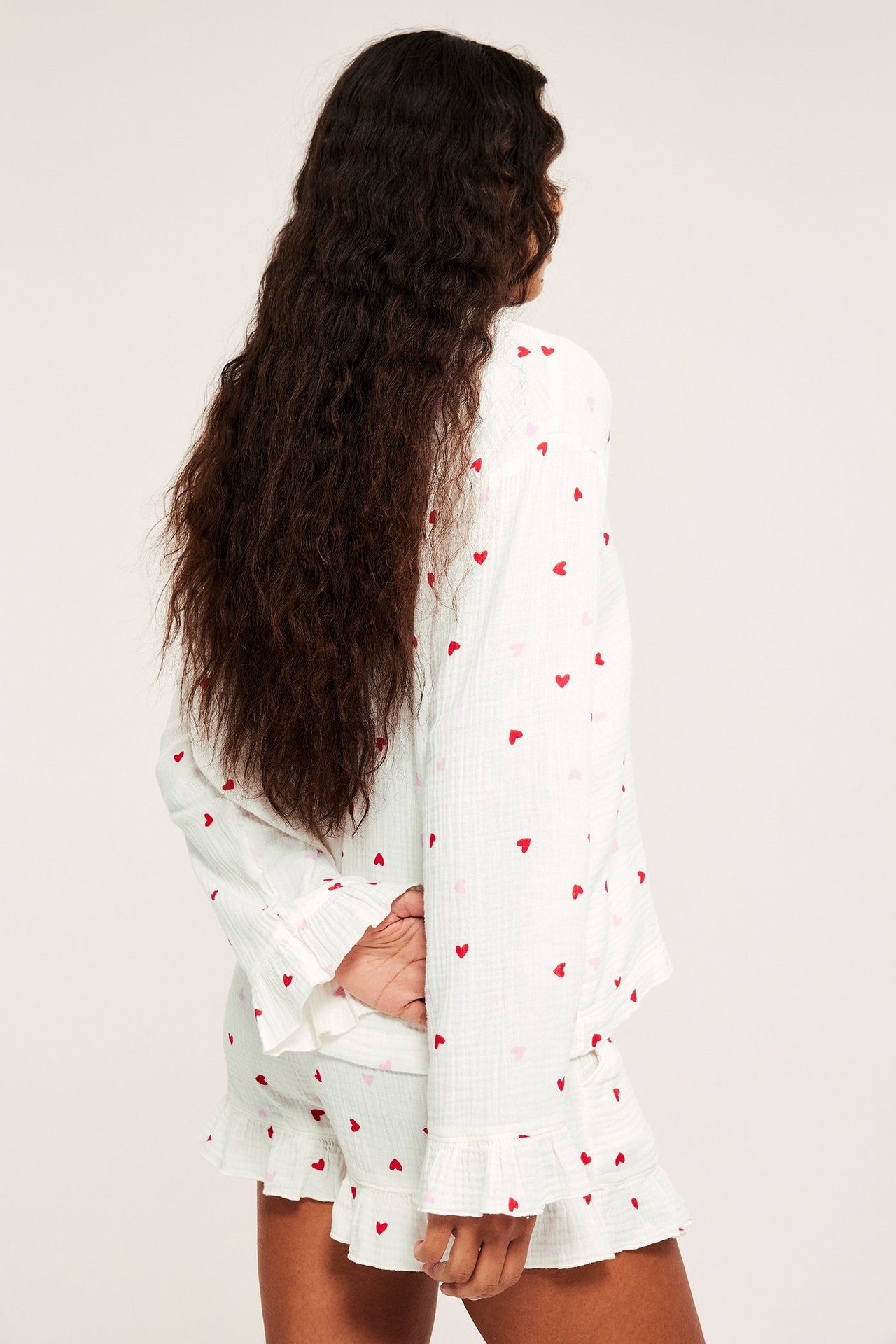 Naisen selkäpuoli: valkoinen puuvillapyjamapaita punaisilla sydämillä, röyhelöillä ja shortseilla.