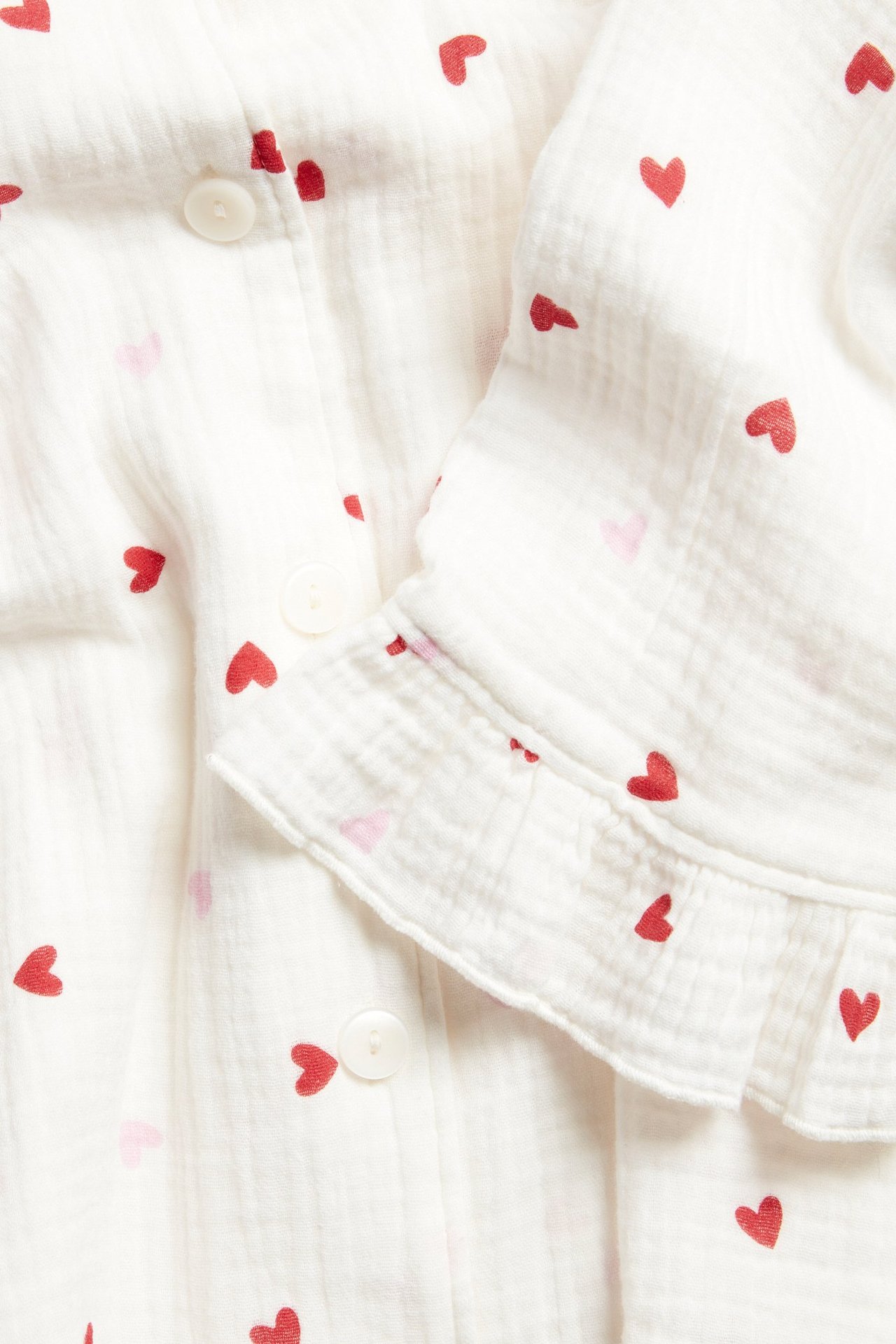 Lähikuva valkoisesta puuvillapyjamapaidasta pinkillä sydänkuviolla, napeilla ja röyhelöhihansuilla.