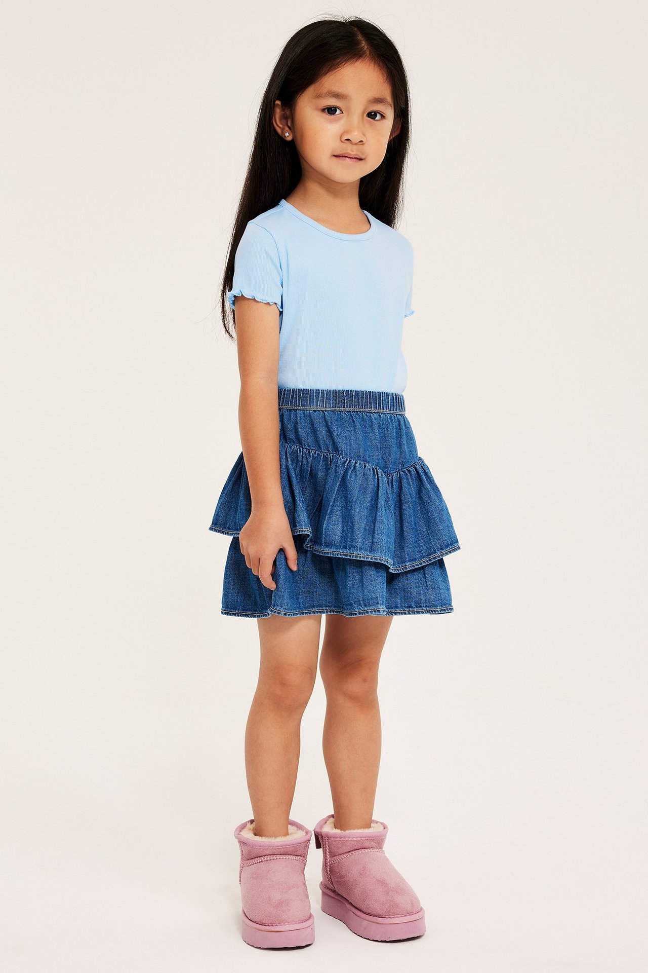 Front view: Kid bär en ljusblå ribbad kortärmad topp, blå jeanskjol med volanger, och rosa stövlar.