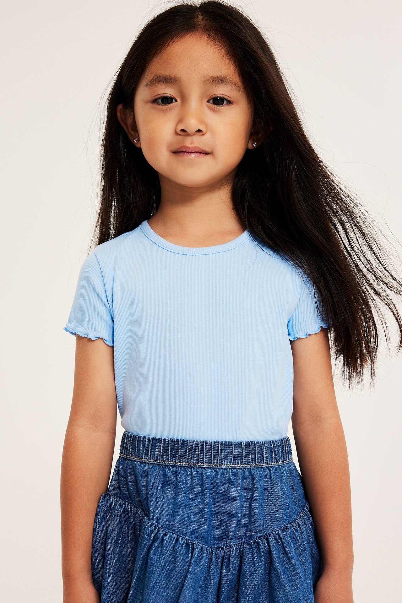 Frontvy av ett barn i en ljusblå ribbad kortärmad topp med babylockkanter och en jeanskjol.