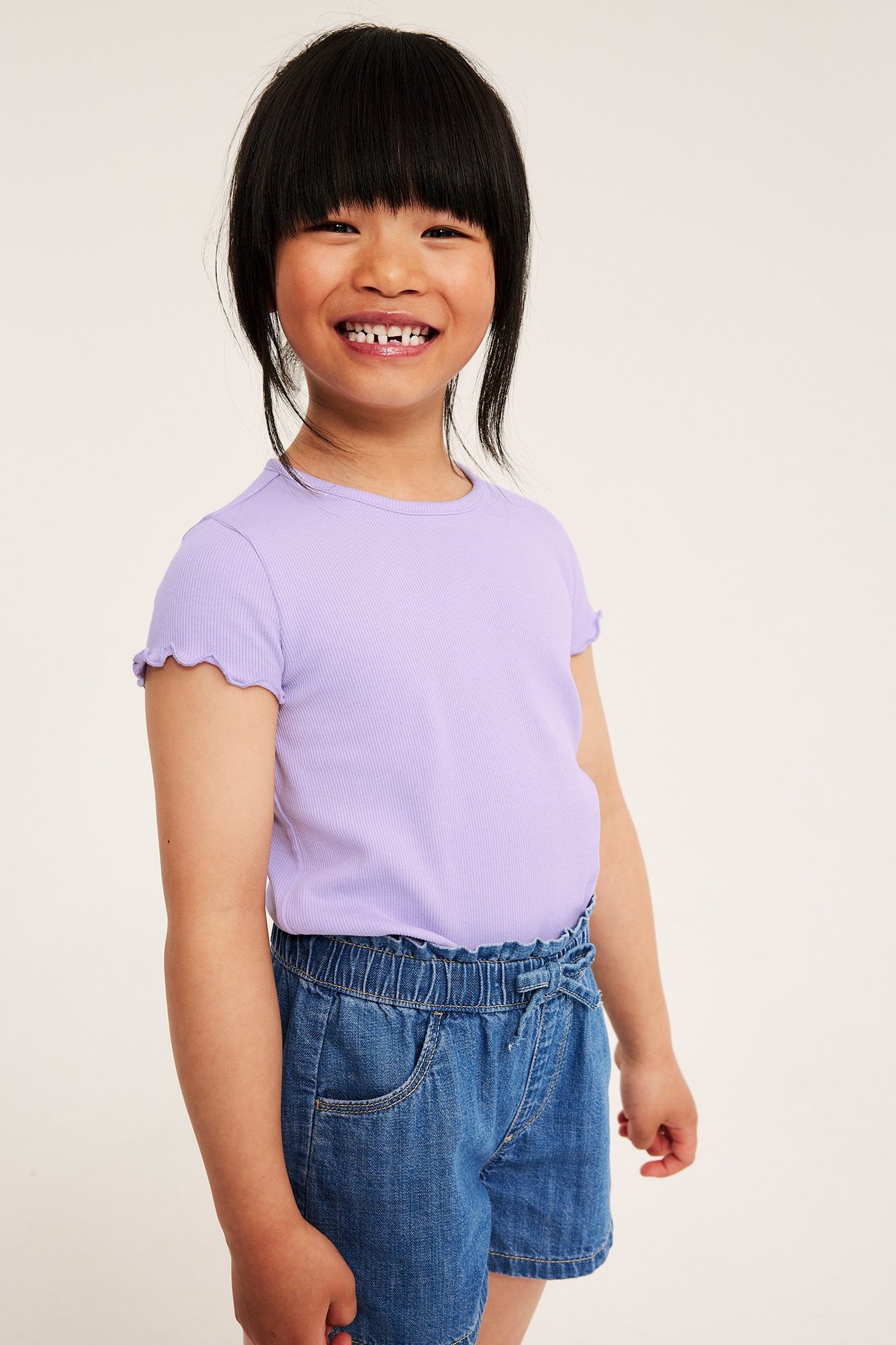 Barn iført en lyslilla ribbet kortarmet topp med babylock-kanter og blå denimshorts.