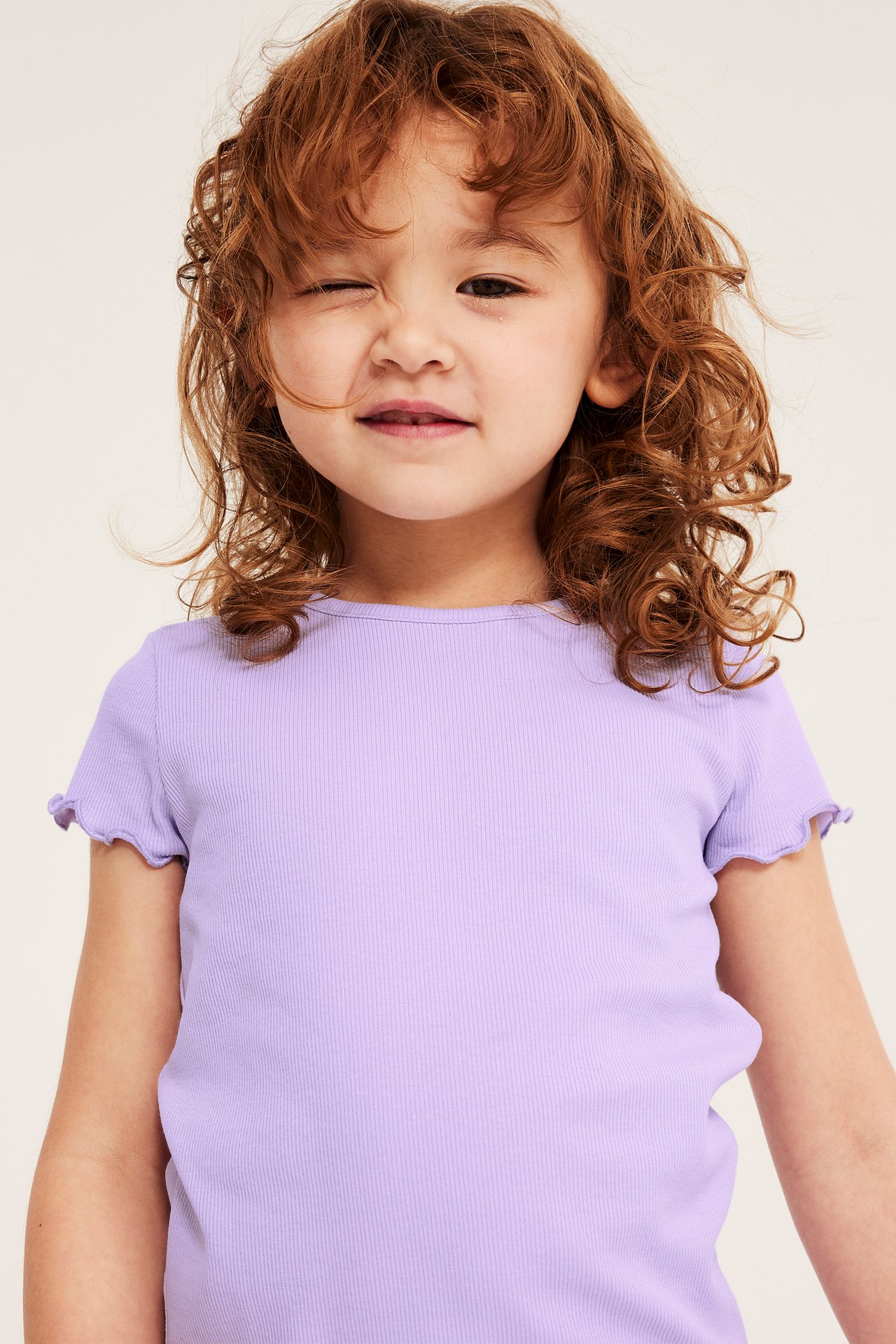 Lähikuva edestä lapsesta, jolla on vaaleanliila ribattu lyhythihainen toppi, jossa on babylock-reunat.