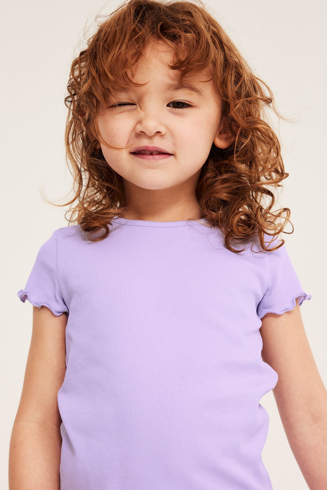 Närbild framifrån på ett barn i en ljuslila ribbad kortärmad topp med babylocksömmar.