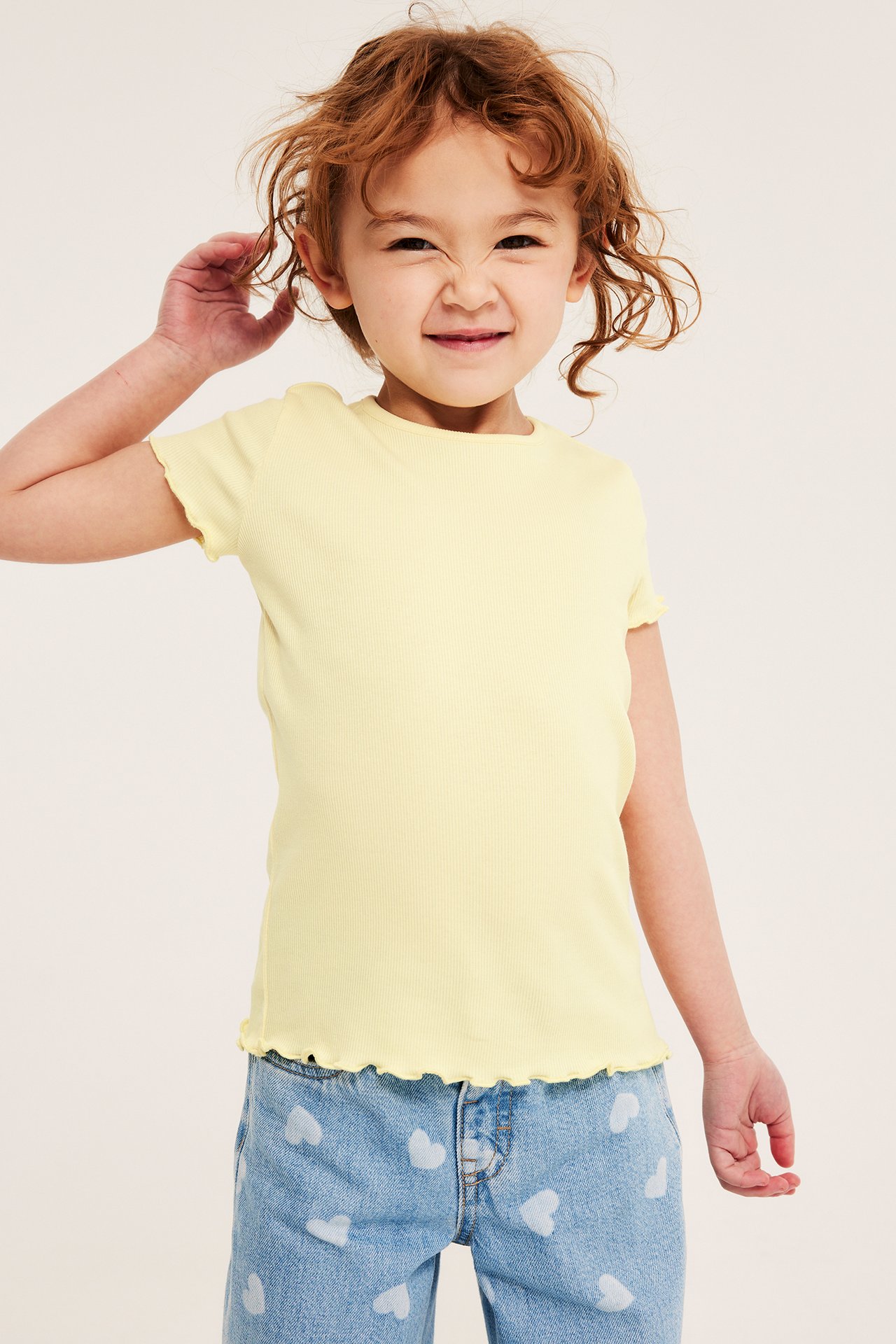 Edestäpäin näkymä lapsesta vaaleankeltaisessa ribatussa lyhythihaisessa topissa ja sinisissä sydänfarkuissa.