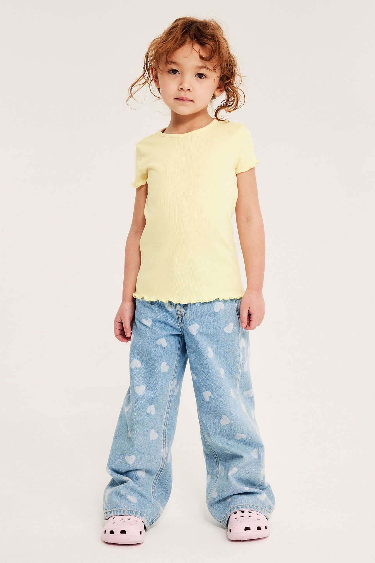 Lapsi edestä: vaaleankeltainen ribbitoppi, siniset sydänfarkut ja vaaleanpunaiset pistokkaat.