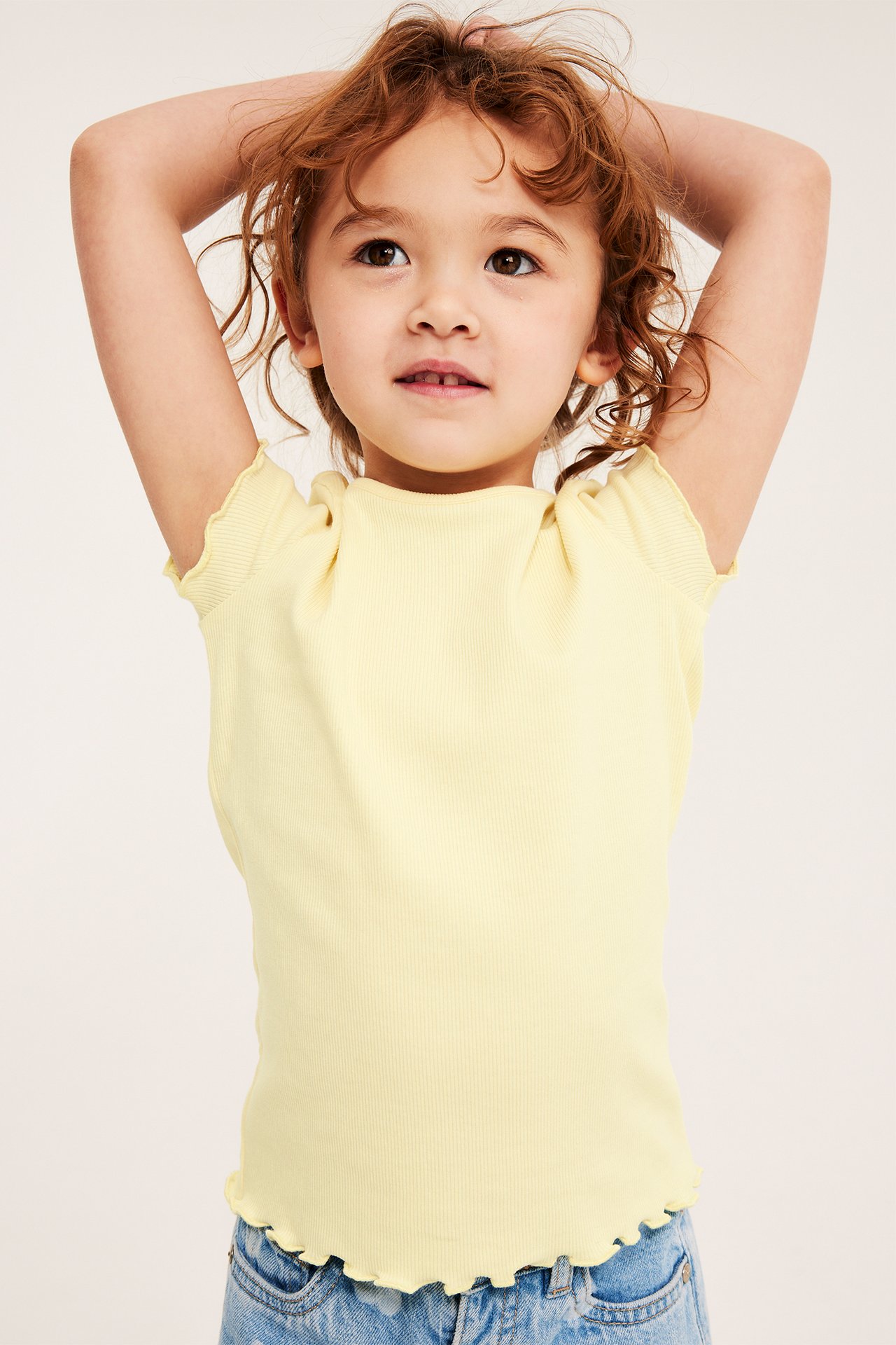 Edestä: lapsi yllään vaaleankeltainen ribbitoppi babylock-reunoilla ja vaaleansiniset farkut.