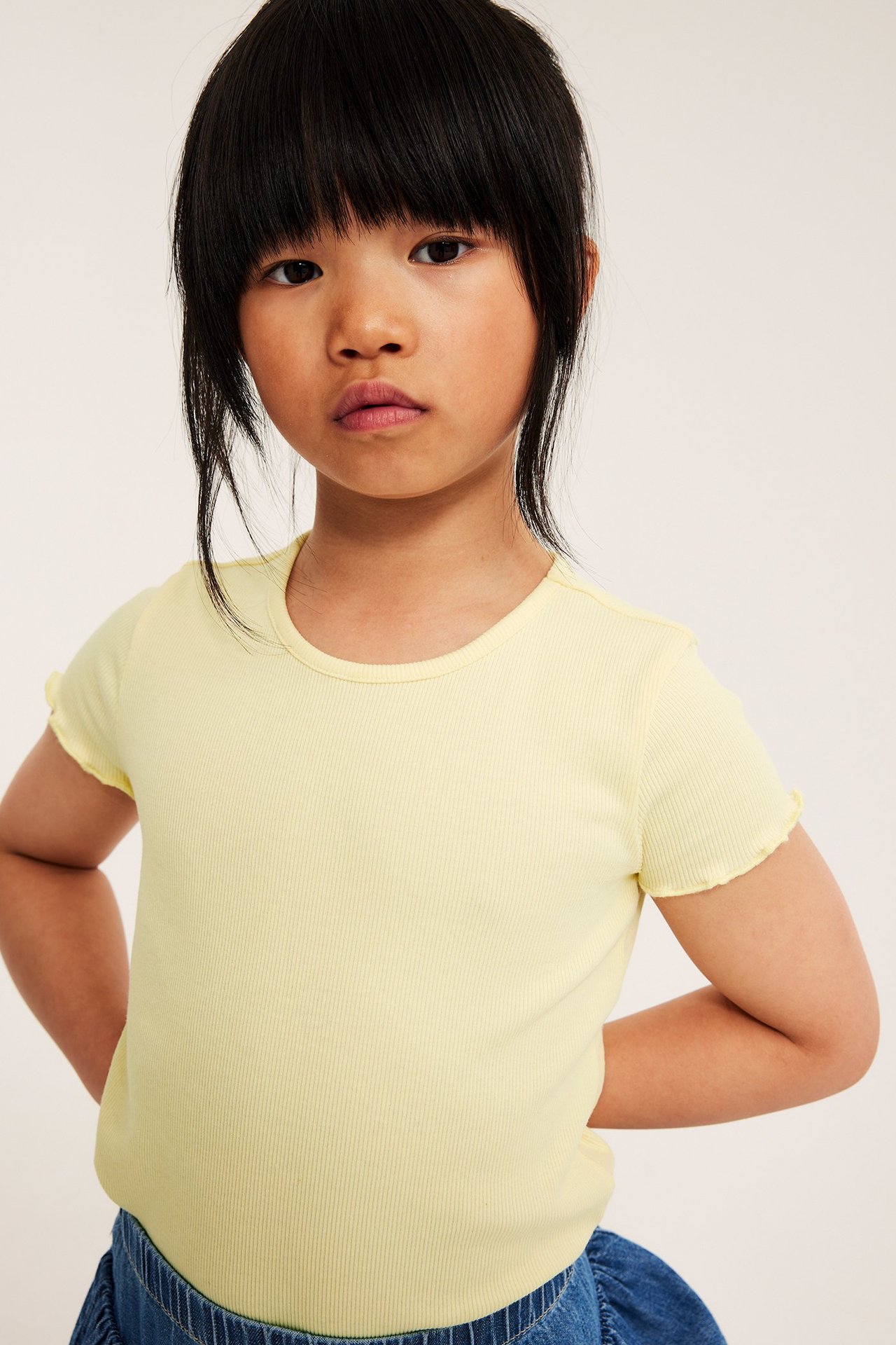 Widok z przodu: dziecko w jasnożółtym prążkowanym topie z krótkim rękawem z brzegami babylock i niebieską spódnicą.