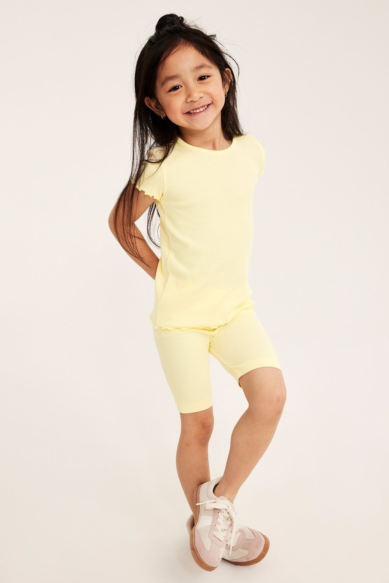 Framifrån: Ett barn i ljusgul ribbad kortärmad topp med babylockkant, matchande shorts och sneakers.