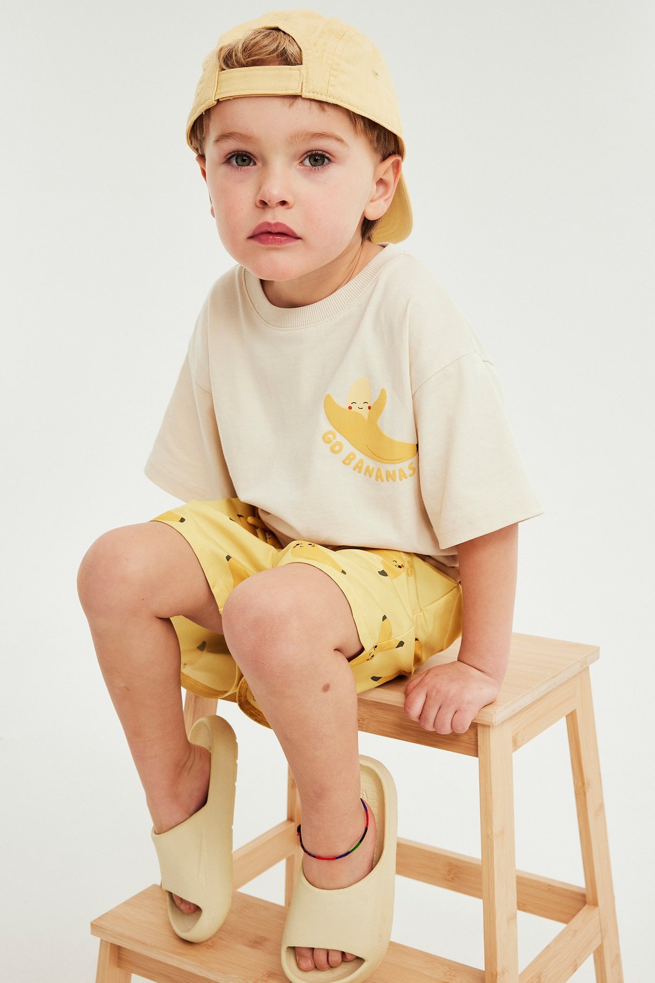 Edestä kuvattu lapsi, keltaiset banaanikuviolliset uimashortsit, beige t-paita, keltainen lippalakki ja beiget sandaalit.