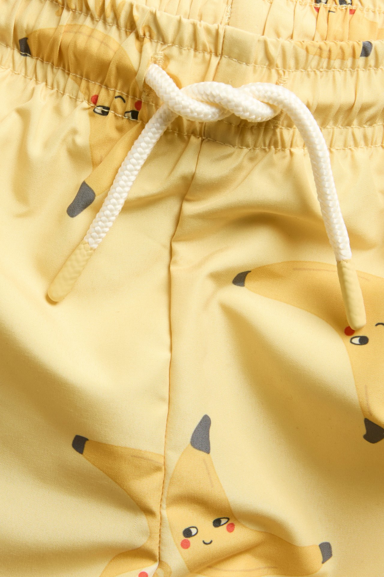 Lähikuva keltaisista uimashortseista banaanikuvioinnilla, resori vyötäröllä ja valkoisella kiristysnyörillä.