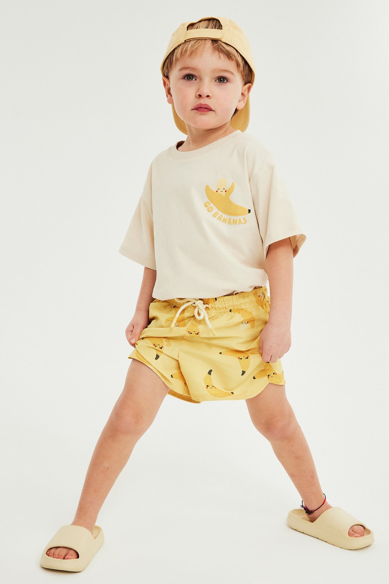 Etunäkymä lapsesta keltaisissa banaanikuvioisissa uimahousuissa, beigessä t-paidassa, lippiksessä ja sandaaleissa.