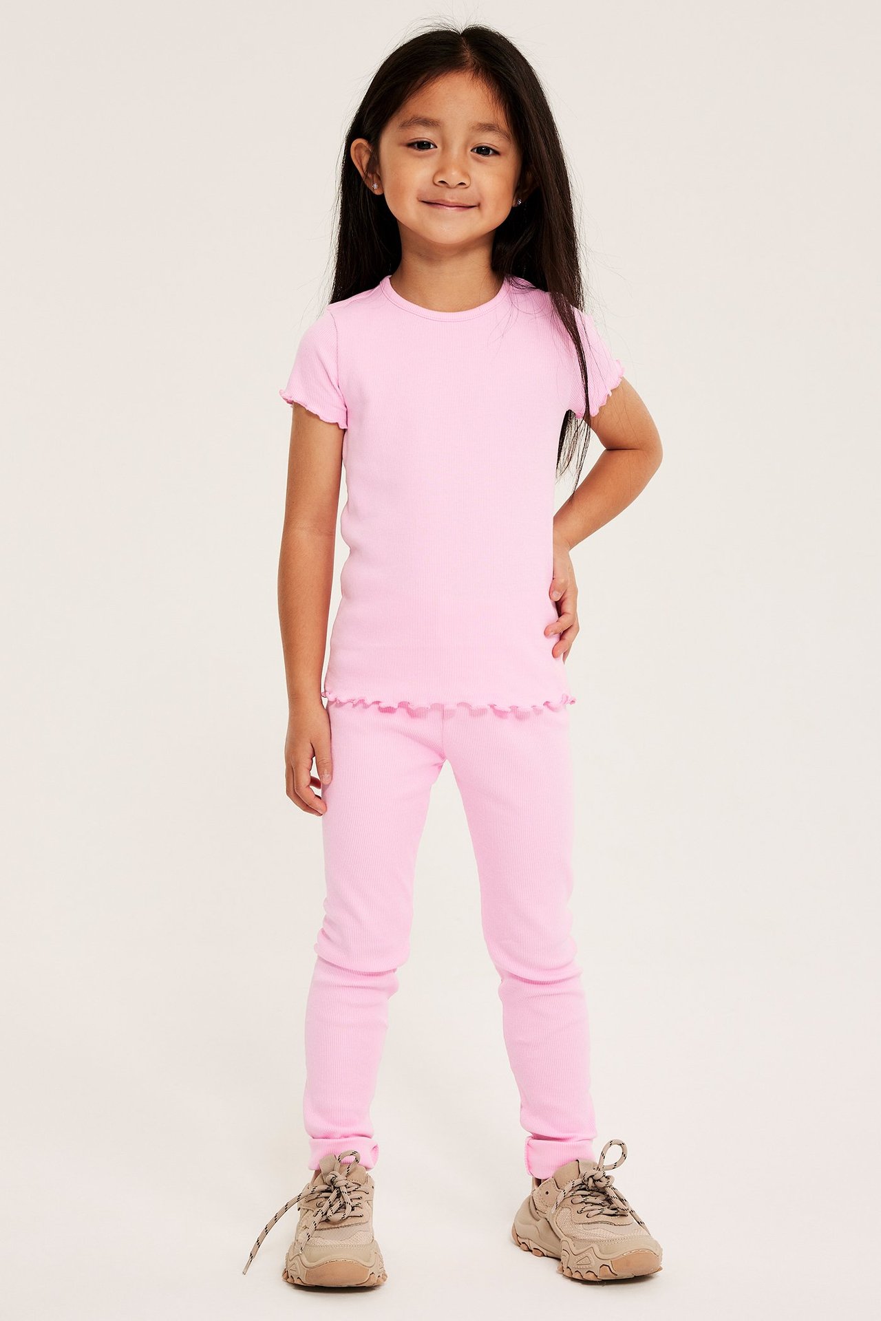 Helkroppsbild framifrån på en tjej som bär ljusrosa ribbade leggings, en matchande kortärmad topp och beige sneakers.